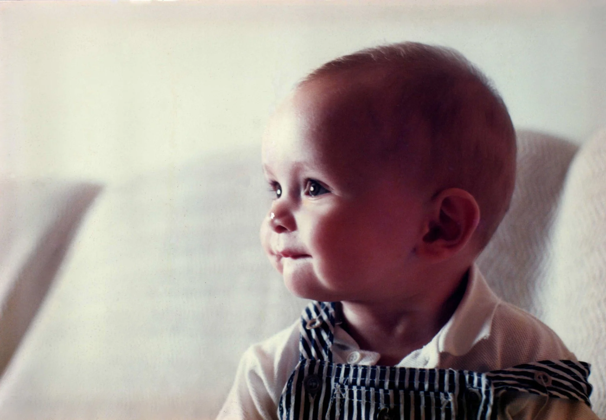 A young child with short hair, wearing a white shirt with a blue and white striped collar, appears to be looking at or reaching for something out of the frame. The background is plain and light-colored.