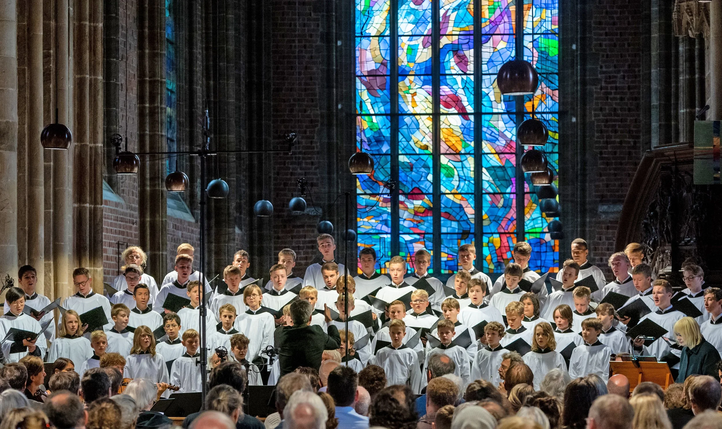 A concert choir of young boys and teenagers dressed in white robes, singing on stage inside a church with a large colorful stained glass window behind them. The audience is seated in front of them, and choir conductor is visible in the foreground.