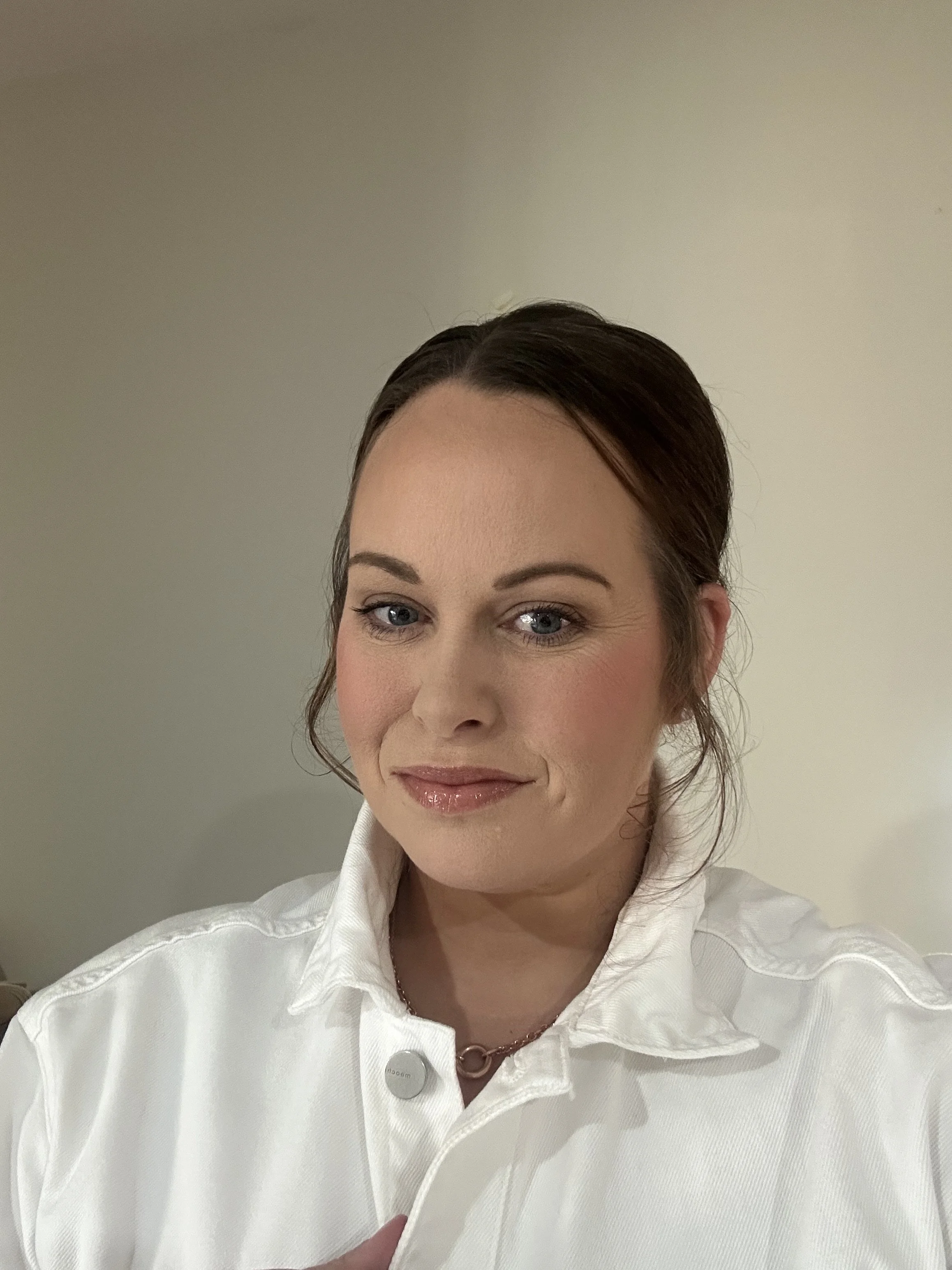 A woman with brown hair and blue eyes wearing a white collared shirt, smiling softly against a plain light-colored background.