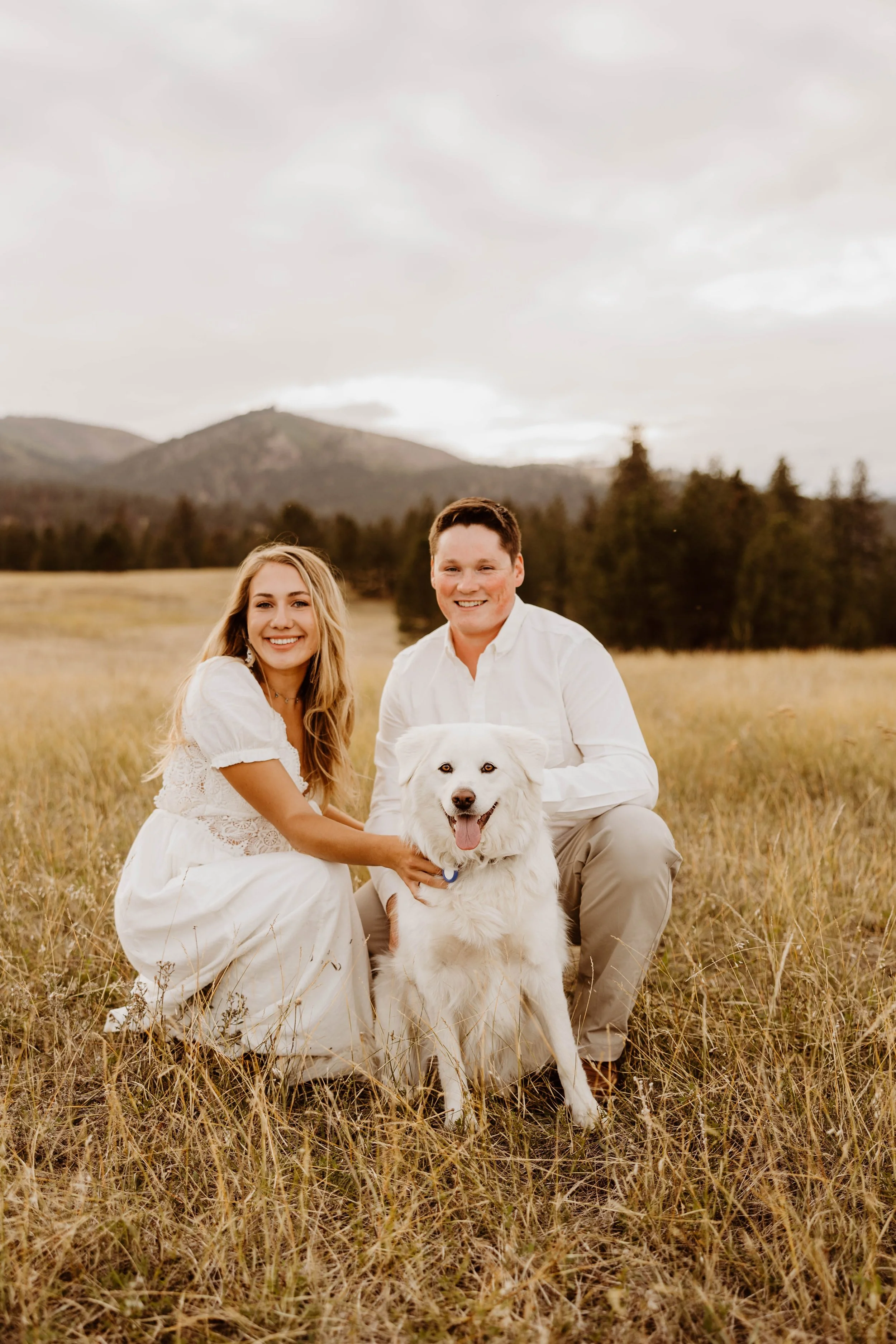 A smiling couple and a happy white dog sitting on a grassy field with mountains and trees in the background.