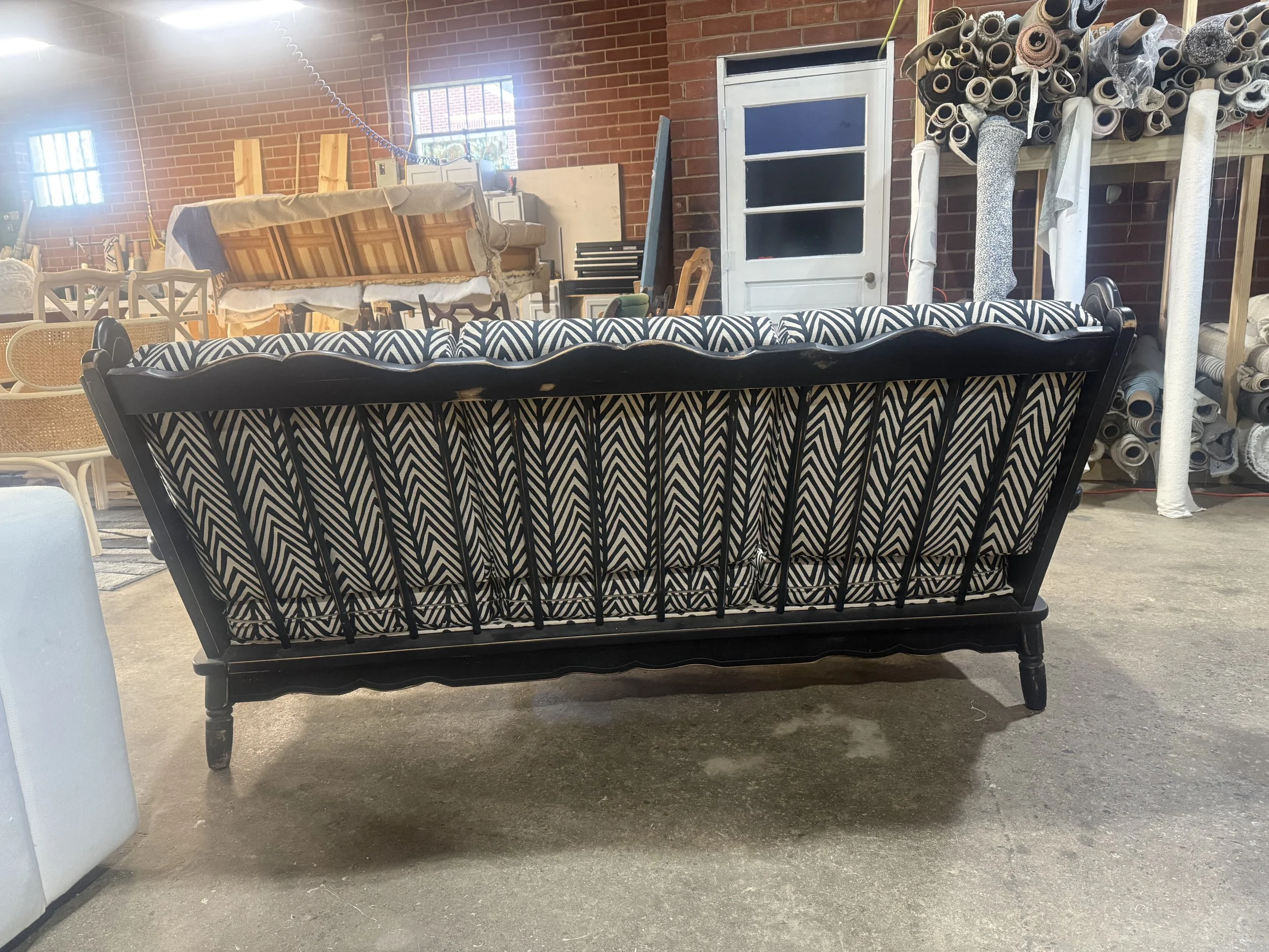 A black and white patterned wooden bench in a storage room with a brick wall. Behind the bench, there are rolls of fabric, wood furniture, and various stored items.