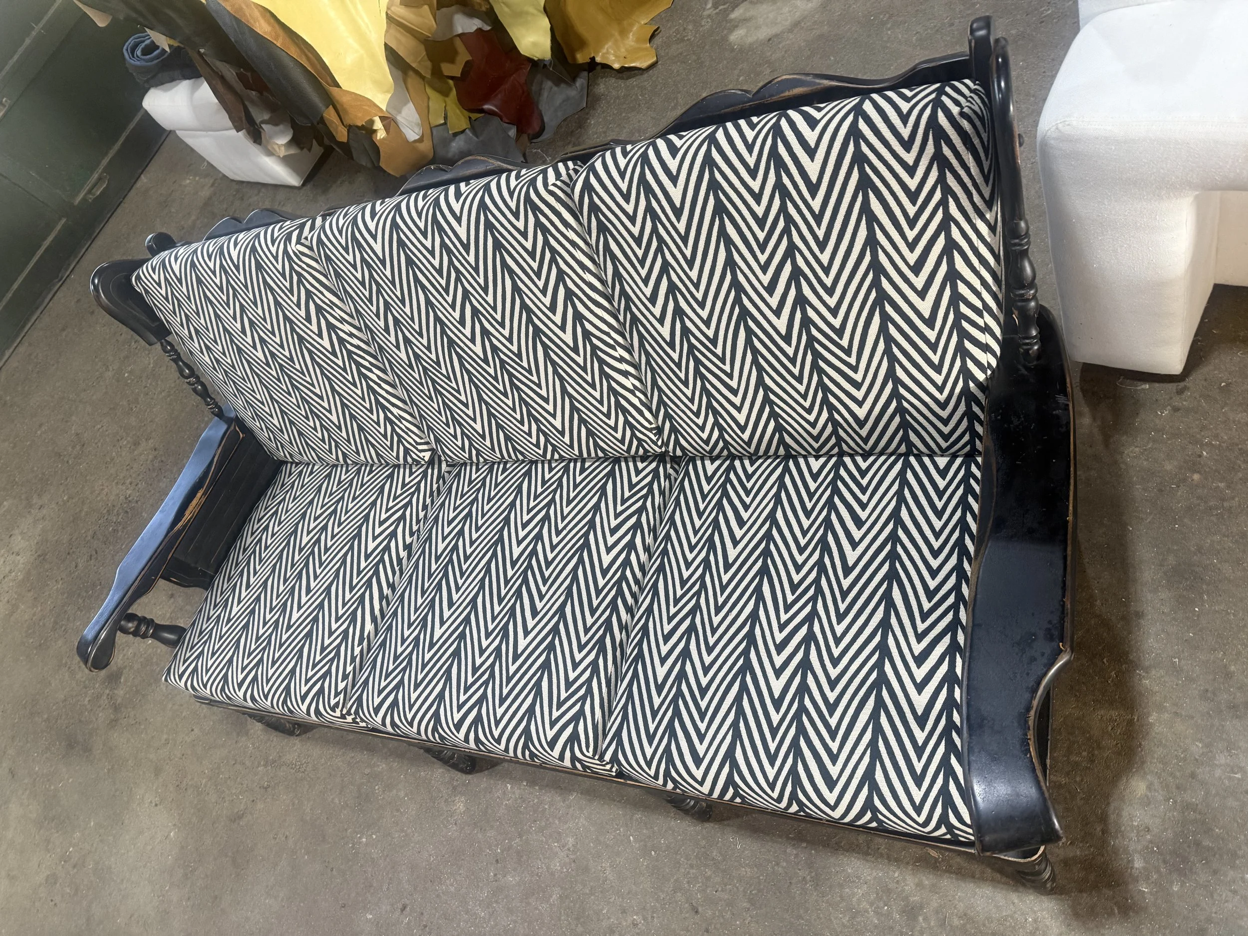A vintage wooden bench with a black and white geometric-patterned cushion or upholstery. The bench is situated on a concrete floor, with various items including fabric or leather scraps and a white bag in the background.