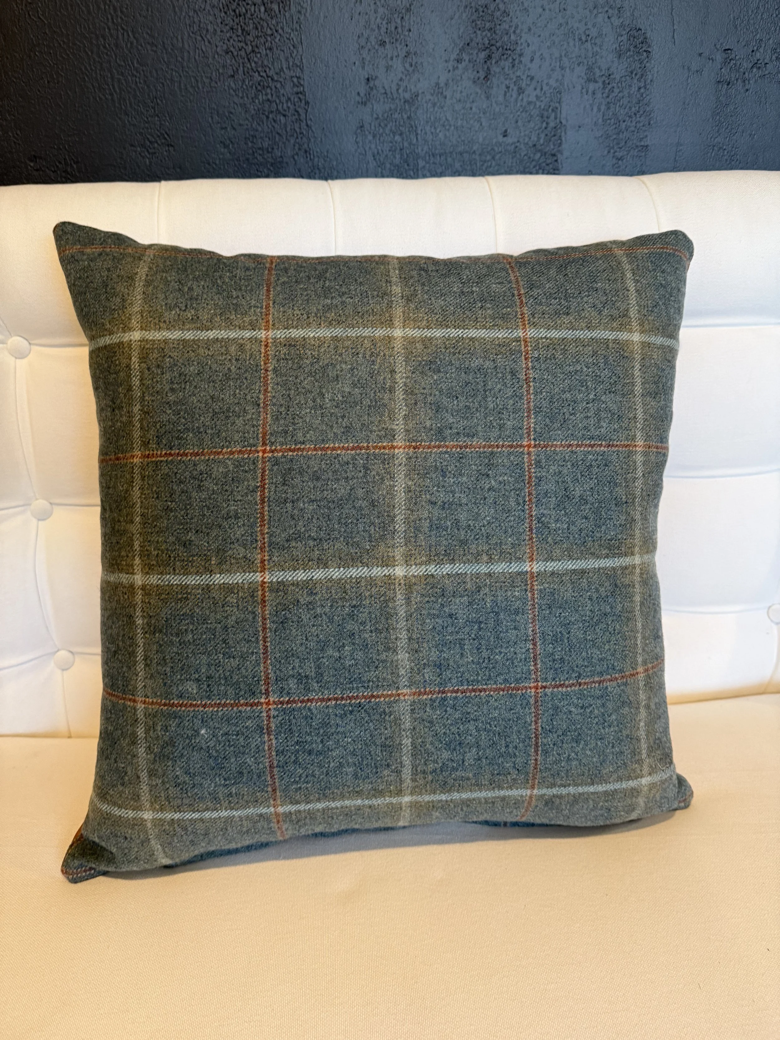 A decorative cushion with a plaid pattern in shades of gray, beige, and red on a cream-colored headboard against a dark blue wall.