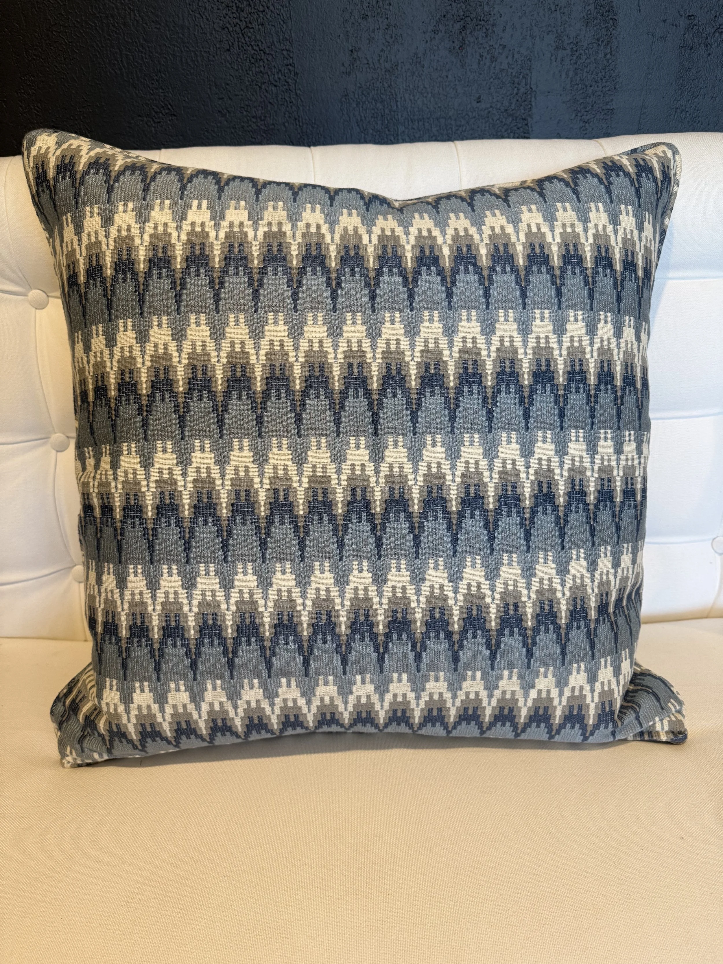 A decorative pillow with a geometric pattern in shades of grey, black, and cream, placed on a white tufted headboard against a dark blue wall.