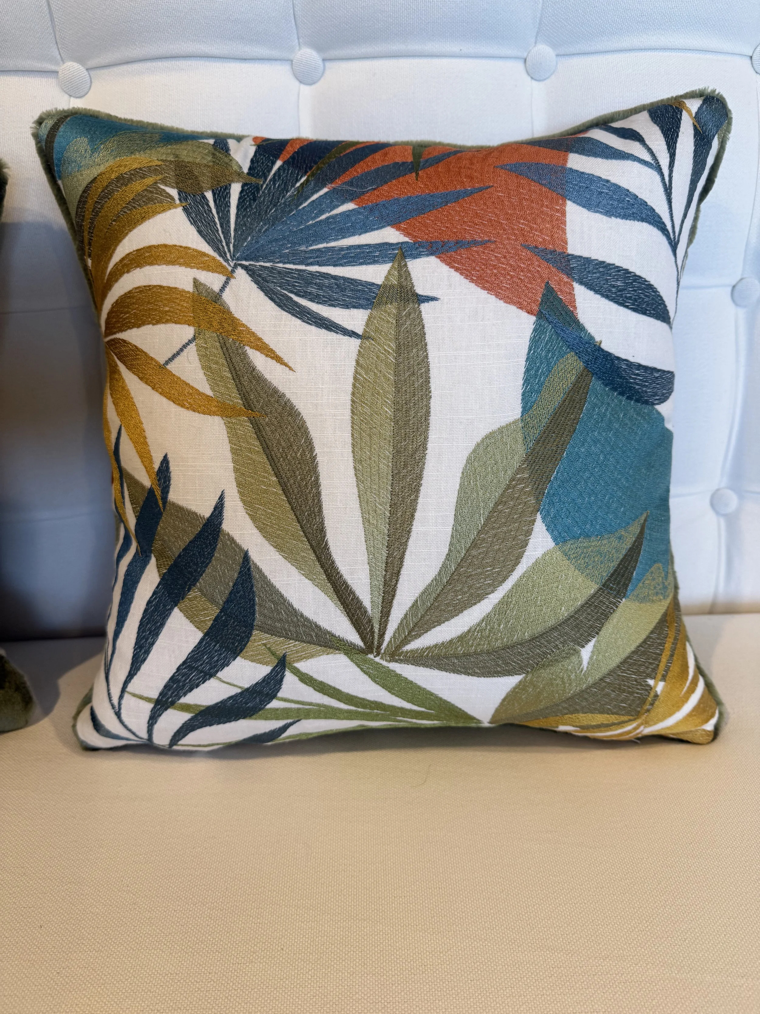 Decorative cushion pillow with a botanical leaf pattern in shades of green, blue, orange, and yellow on a white background, placed on a beige surface against a tufted white headboard.