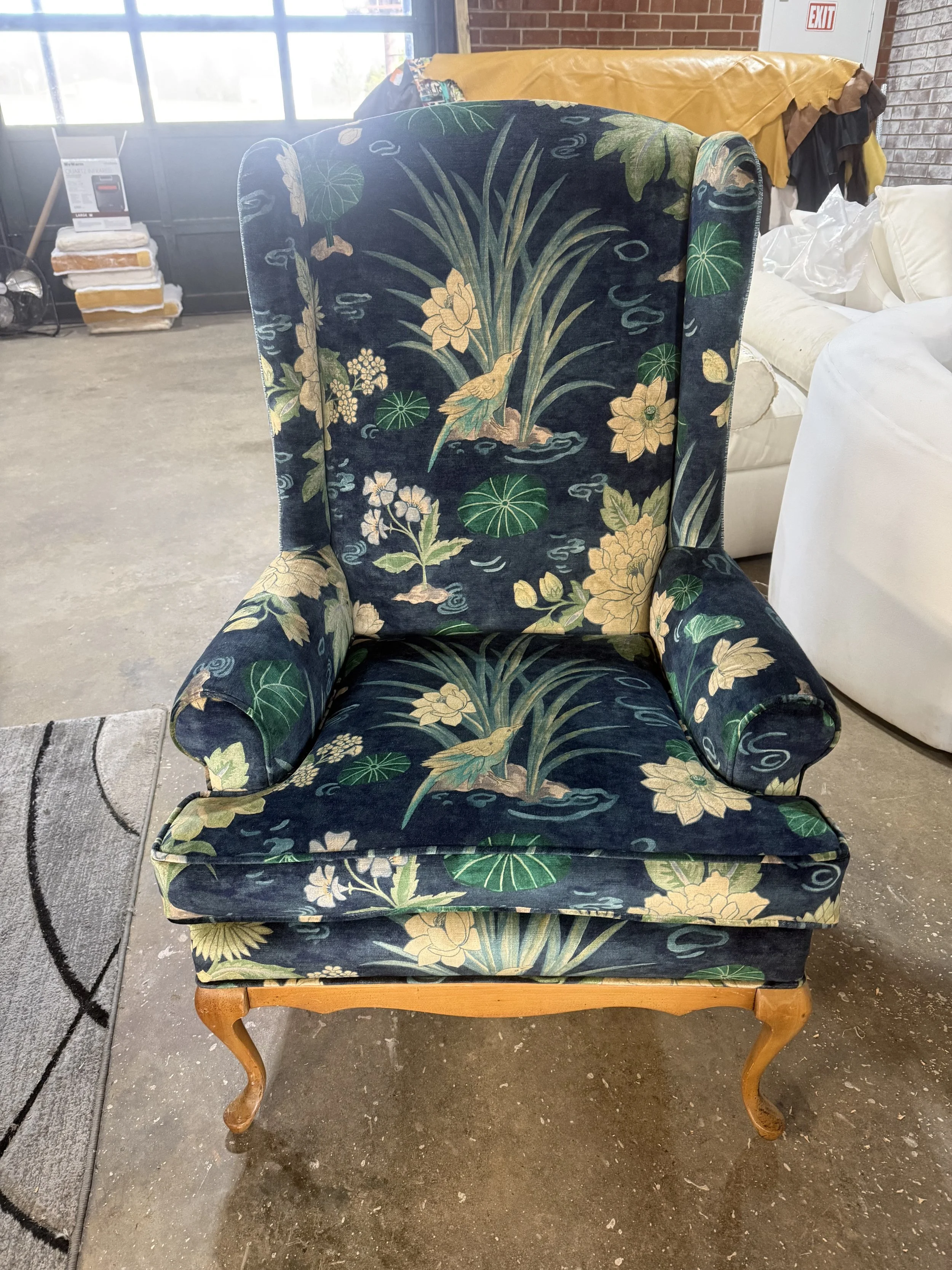 A vintage armchair with a wooden frame and curved legs, upholstered in fabric with a blue background featuring a floral and bird pattern. The chair is placed on a concrete floor next to a gray rug with a black border, in a warehouse or showroom setti