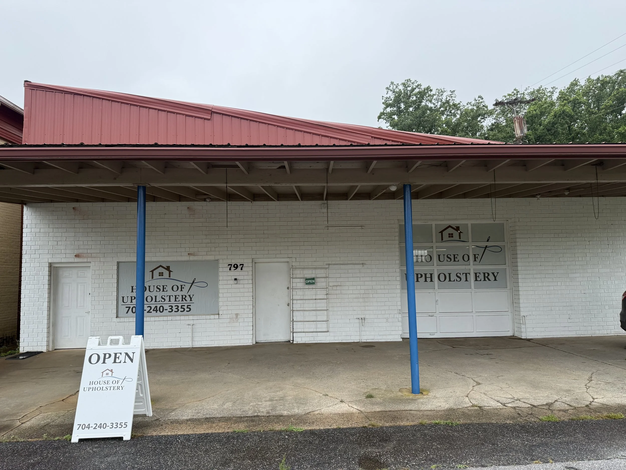The image shows the exterior of a business called 'House of Upholstery' with its signage on the door and a sandwich board sign on the sidewalk that says 'Open.' The building has a white brick wall, a red metal roof, and blue support poles. There is a window with a house icon and contact information. The sky is overcast, and the ground is wet, indicating rainy weather.
