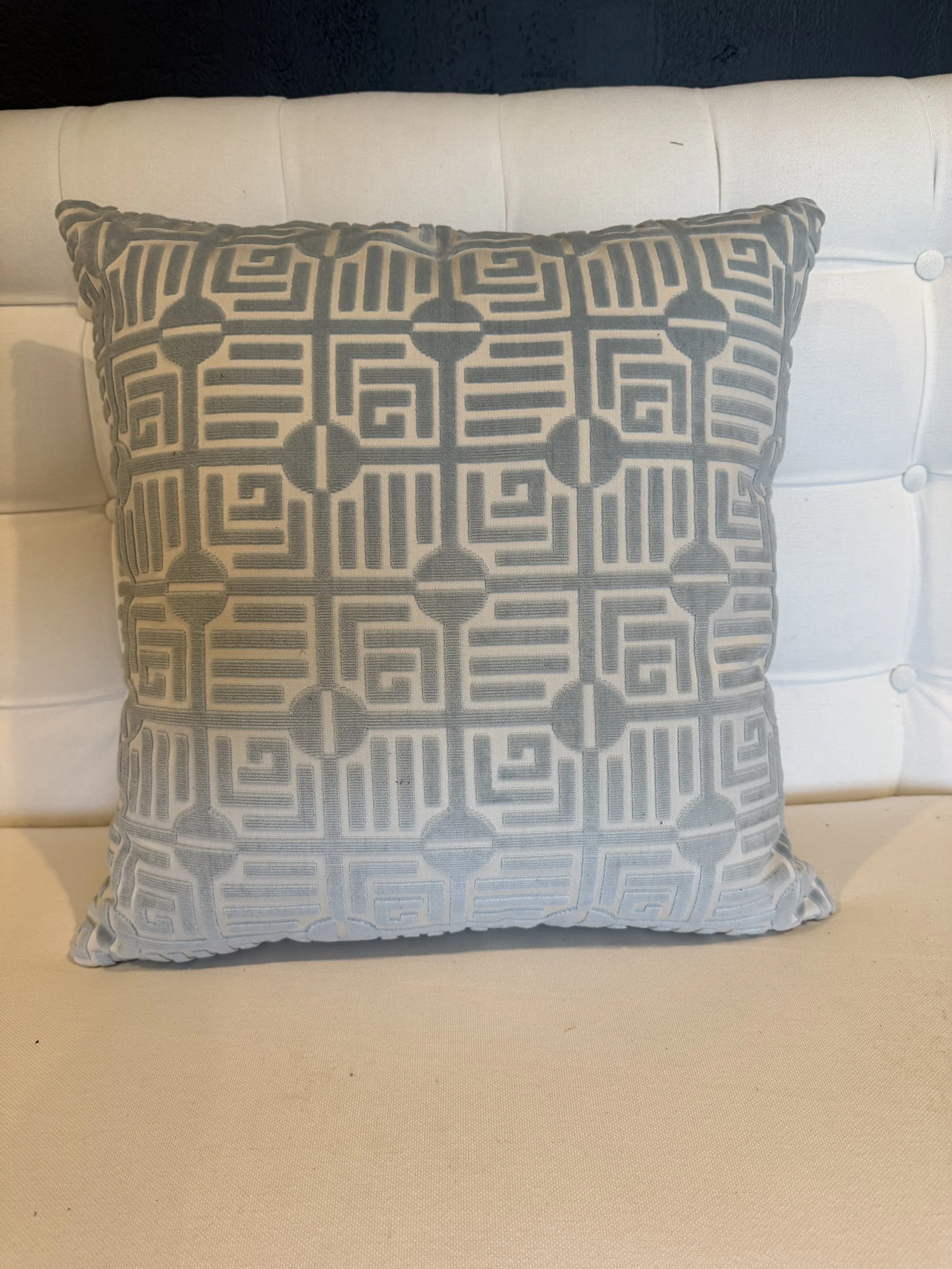 A decorative throw pillow with a geometric pattern in gray and white, placed on a white cushioned headboard of a beige or cream-colored bed.