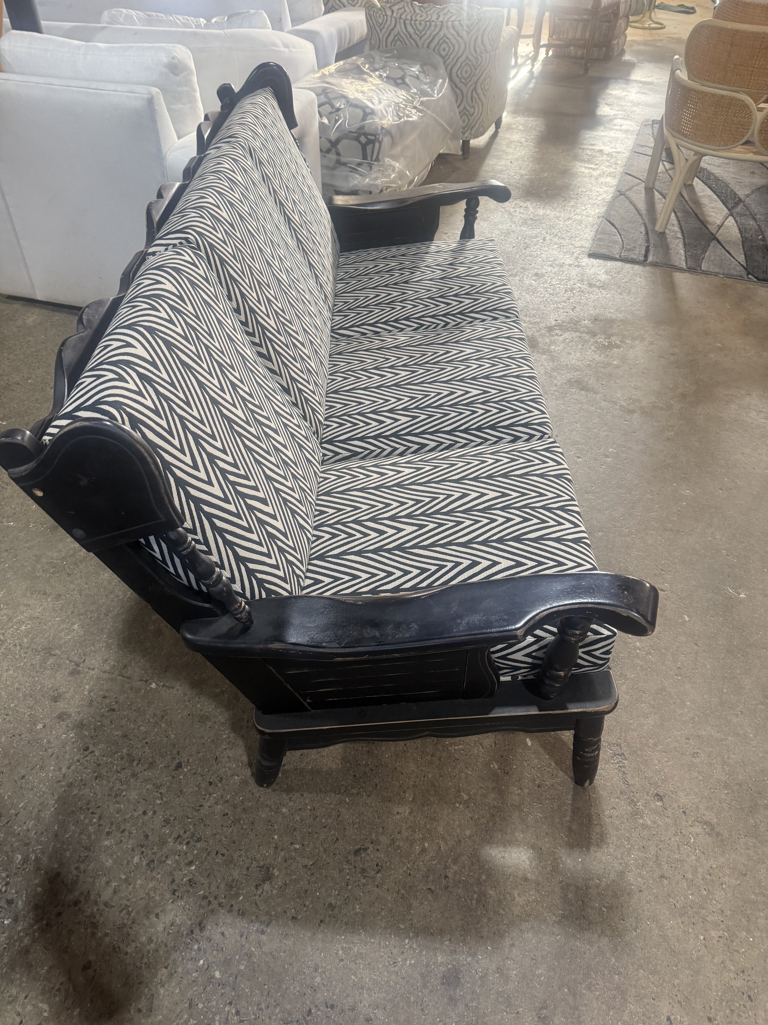 A black wooden bench with a patterned black and white fabric seat and backrest, situated on a concrete floor in a furniture store, with other furniture visible in the background.