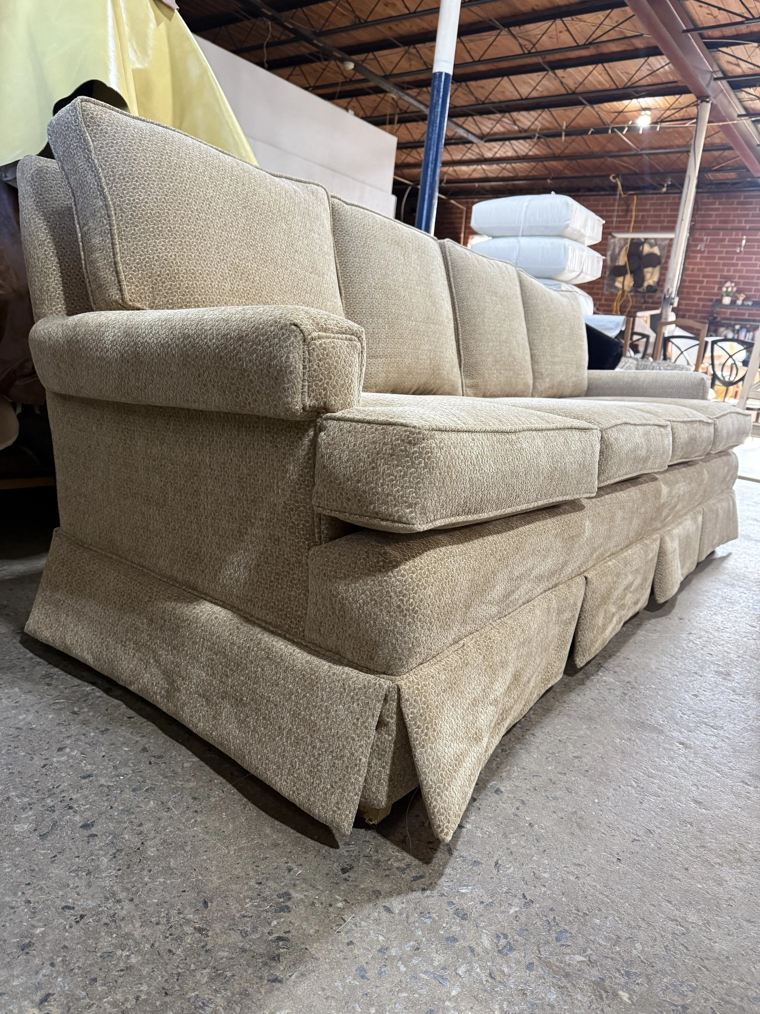 A vintage beige upholstered sofa with a pleated skirt, placed on a concrete floor in a warehouse or furniture store.