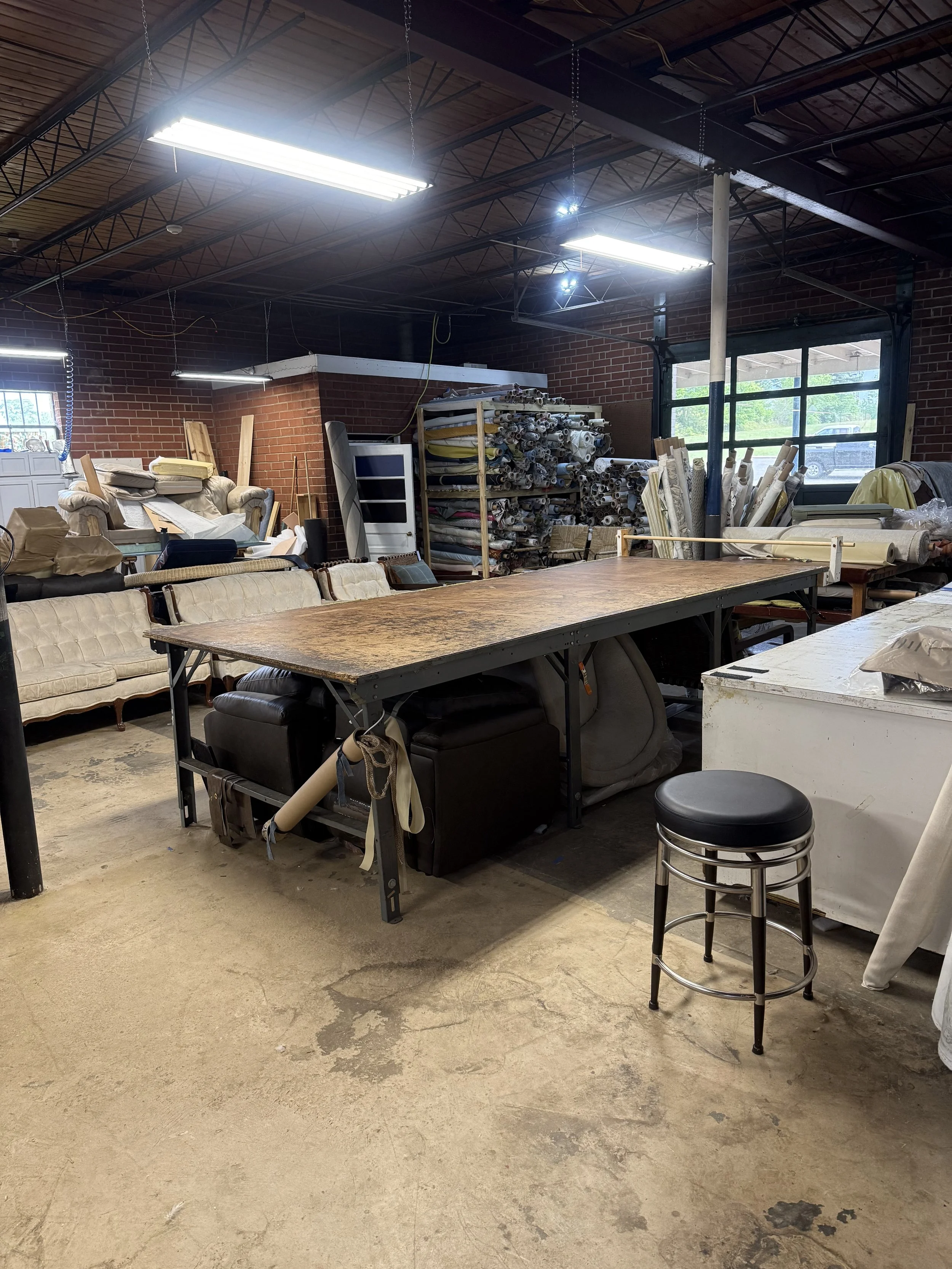 A cluttered workspace inside a building with a large worktable, sofas, storage shelves of rolled fabrics and curtains, a small metal stool, and a large window allowing natural light.