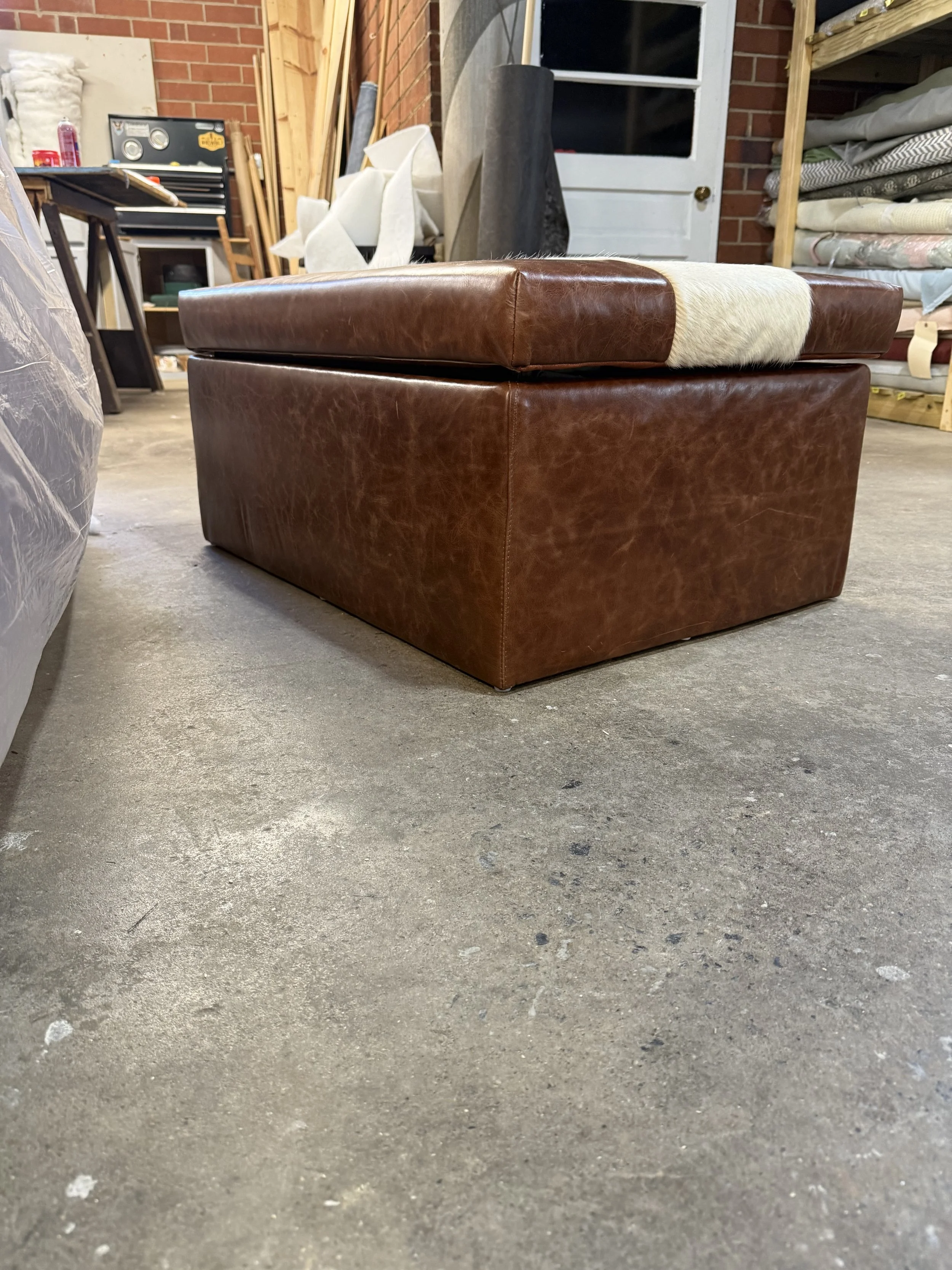 A brown leather ottoman with a white horsehair strip on top, placed on a concrete floor in a workshop or storage room with shelves of fabric and wood in the background.
