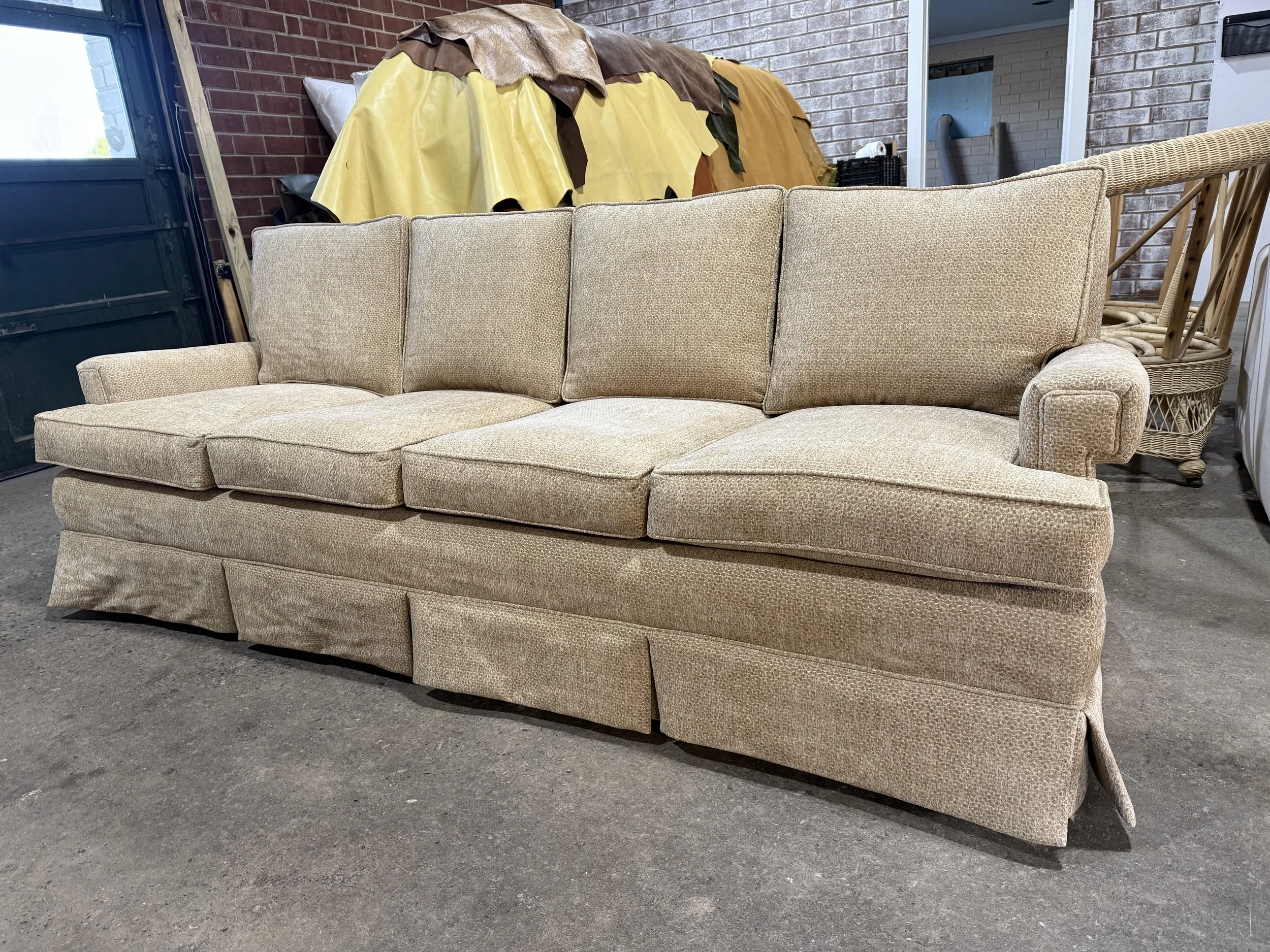 Vintage beige sofa with textured fabric and curved armrests, placed on a concrete floor in a dimly lit room with brick walls and furniture in the background.