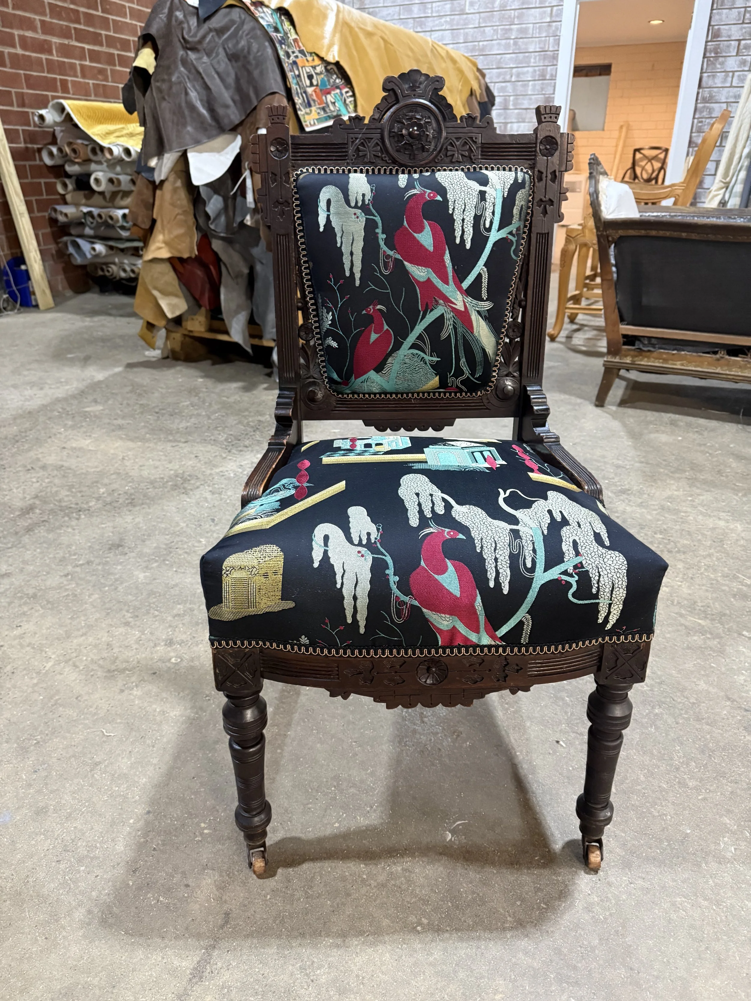 An ornate wooden chair with detailed carvings, upholstered with fabric depicting colorful flamingos and tropical motifs. The chair is placed on a concrete floor, with various items and furniture in the background.