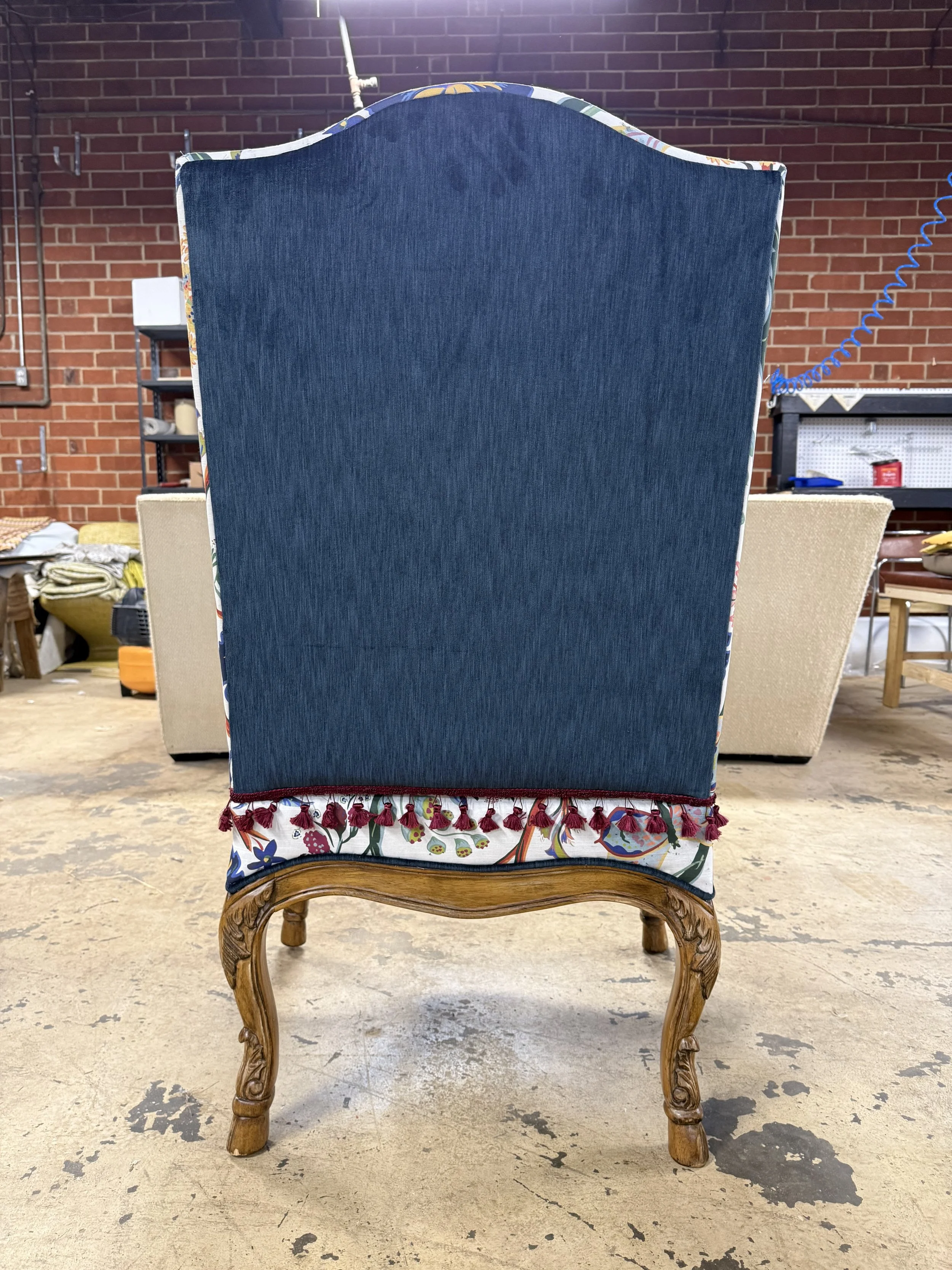 A vintage wooden chair with curved legs, padded seat, and a tall backrest. The backrest is covered in dark blue fabric, while the seat has a colorful floral fabric with tassel trim. The chair is in a workshop or warehouse setting with exposed brick w
