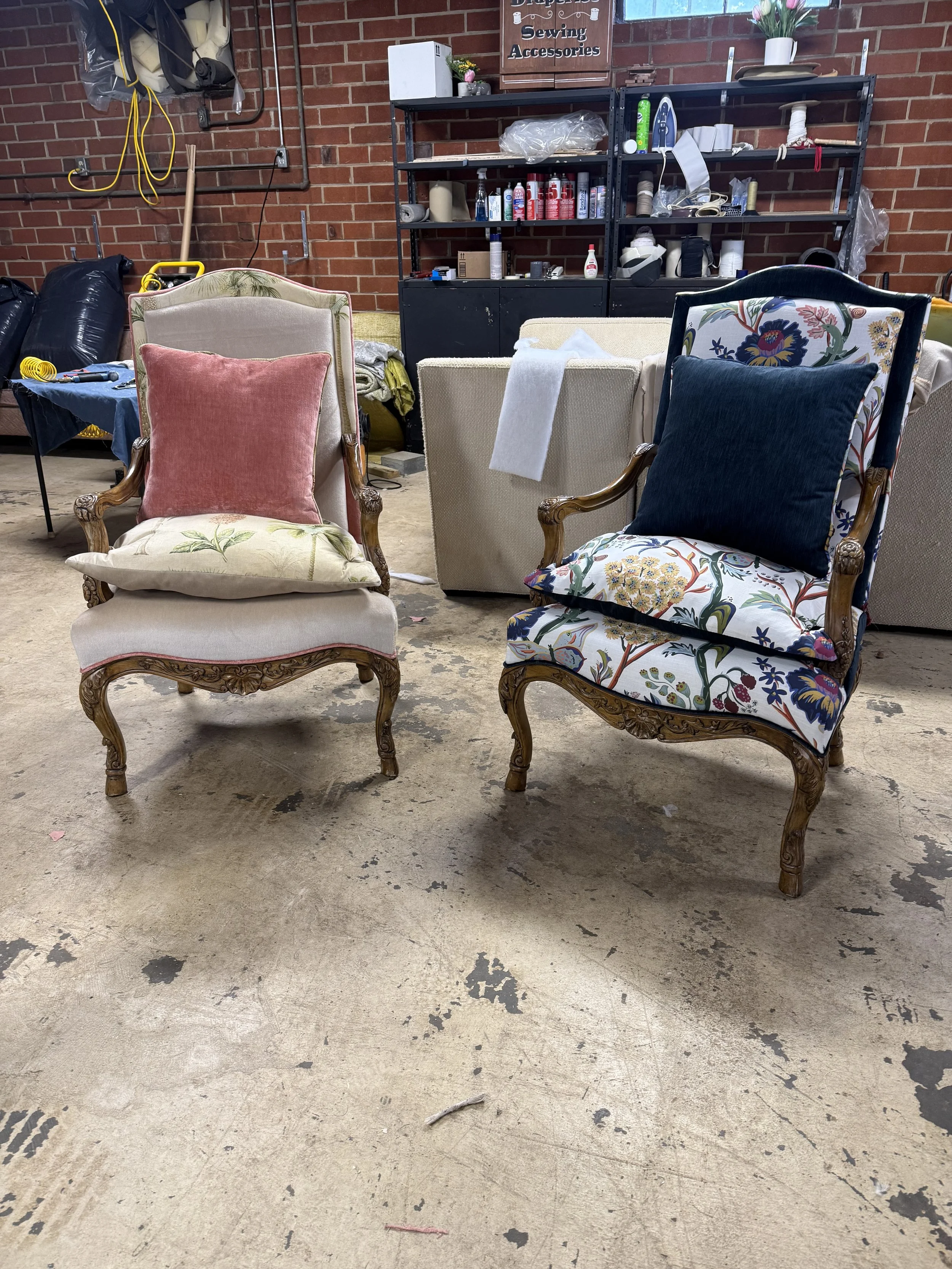 A collection of vintage chairs with decorative pillows is arranged in front of black shelving in a workshop with a brick wall. The floor is concrete and appears worn and stained.