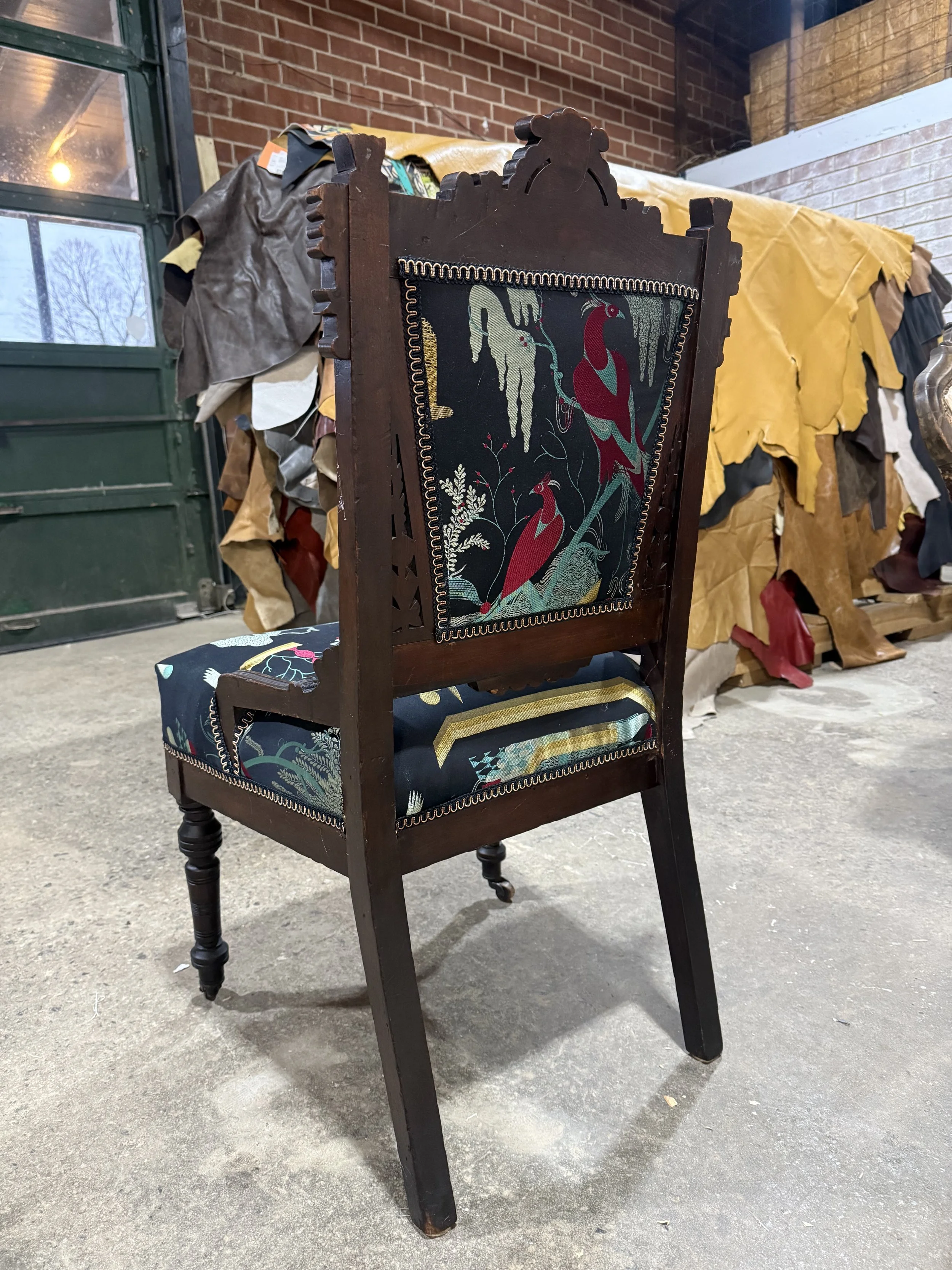 A vintage wooden chair with a carved top and floral upholstery, placed on a concrete floor inside a room with a brick wall and a window.