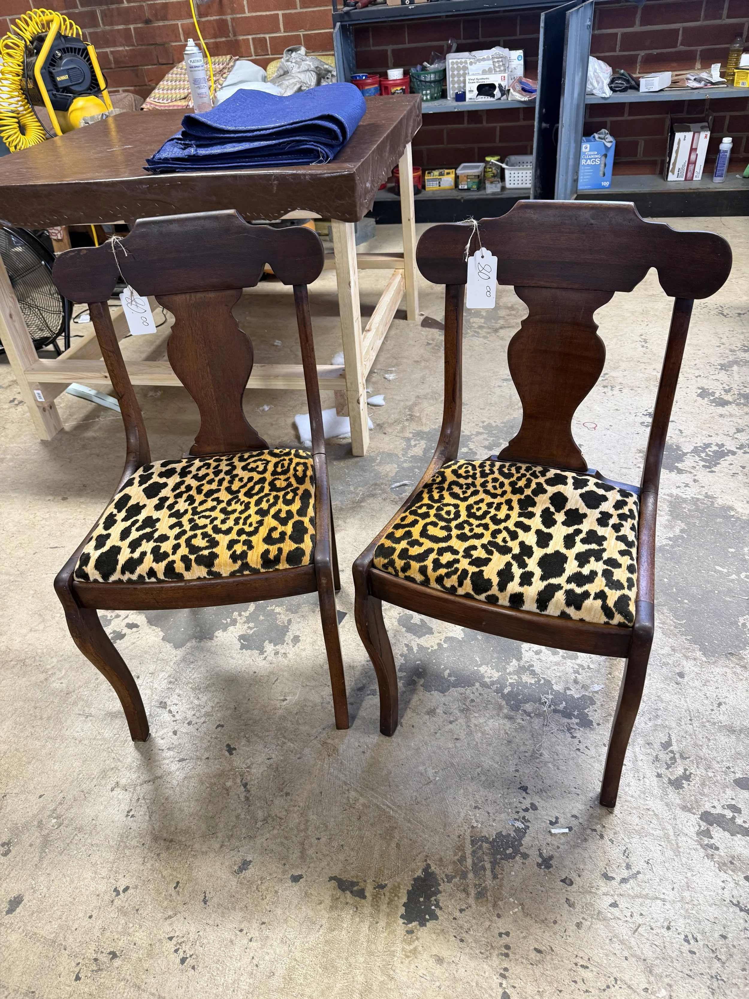 Two wooden chairs with leopard-print upholstered seats in a workshop or flea market setting. A table with folded denim jeans is in the background, along with tools, cleaning supplies, and storage shelves.