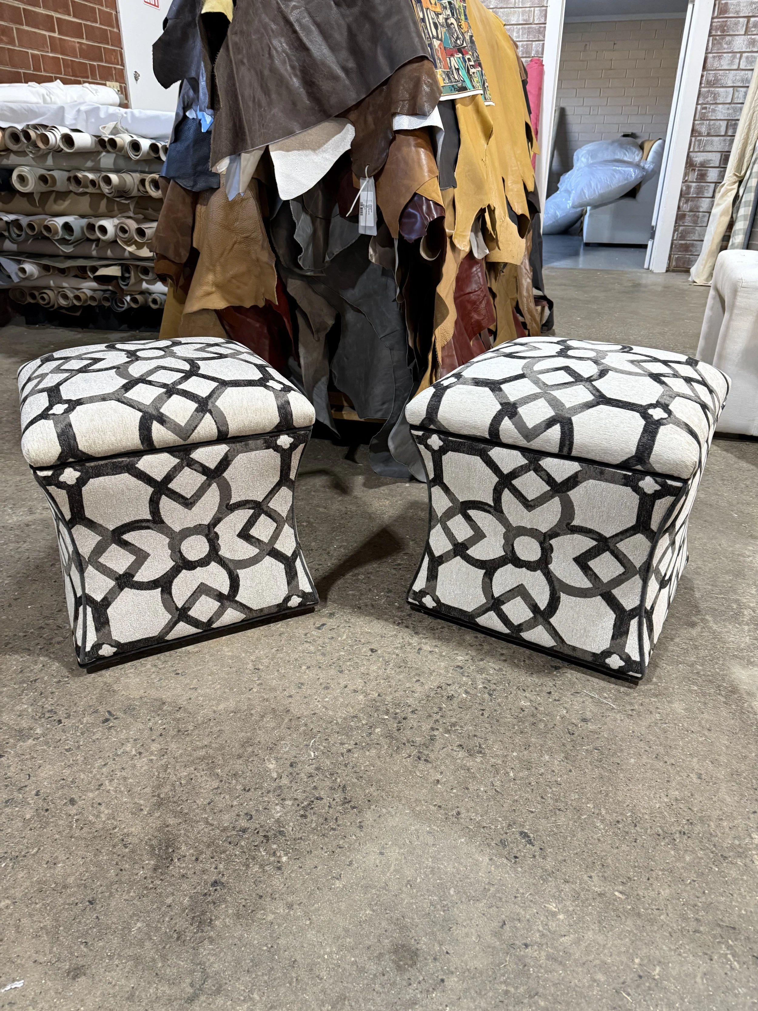 Two upholstered ottomans with black and white geometric pattern are positioned in front of a pile of hanging fabric or leather swatches, with furniture and fabric rolls in the background.