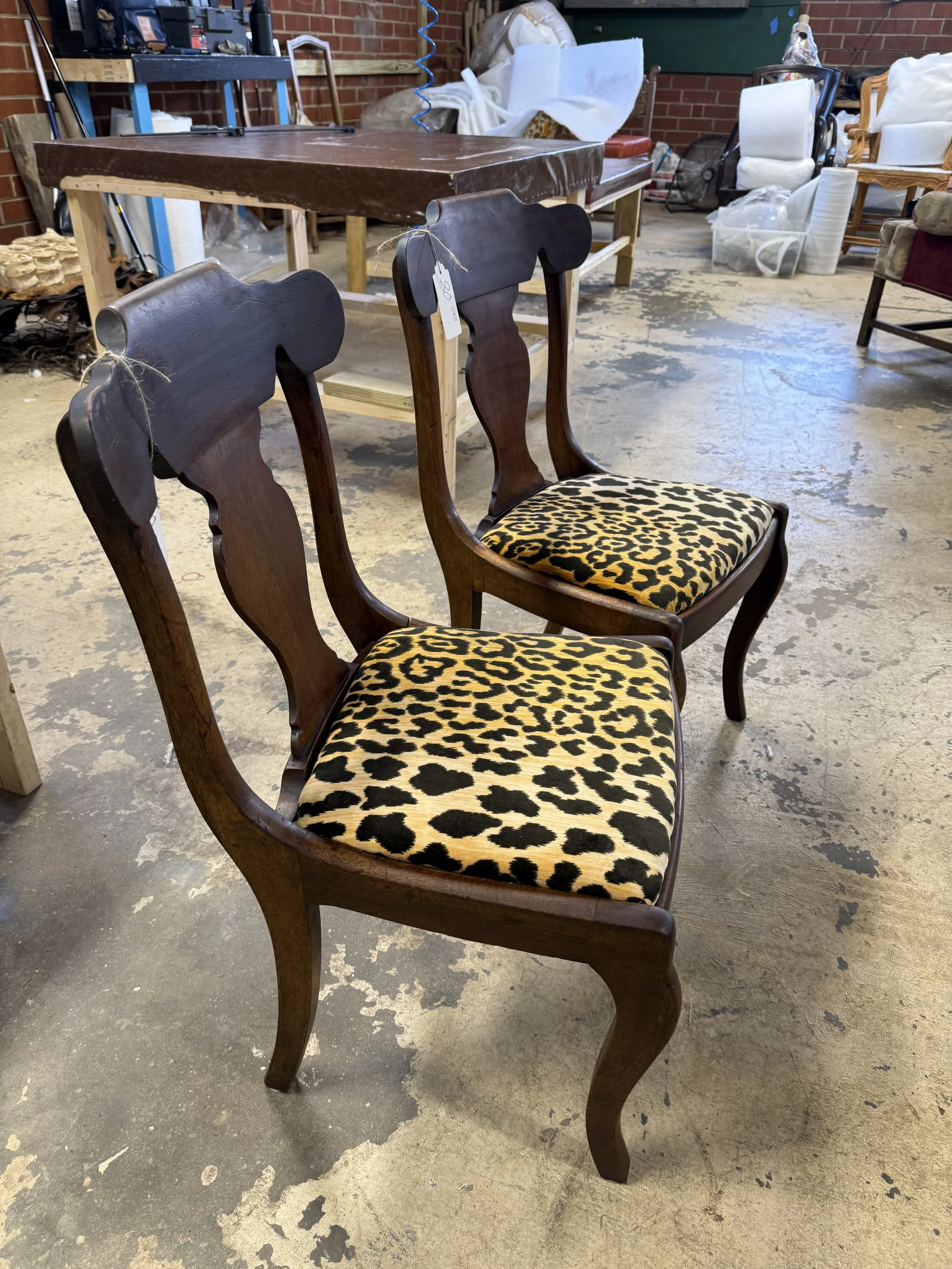 Three wooden chairs with dark brown finish and leopard print cushioned seats are placed in front of a dark brown rectangular table in a cluttered workshop space with brick walls, various furniture, and supplies.