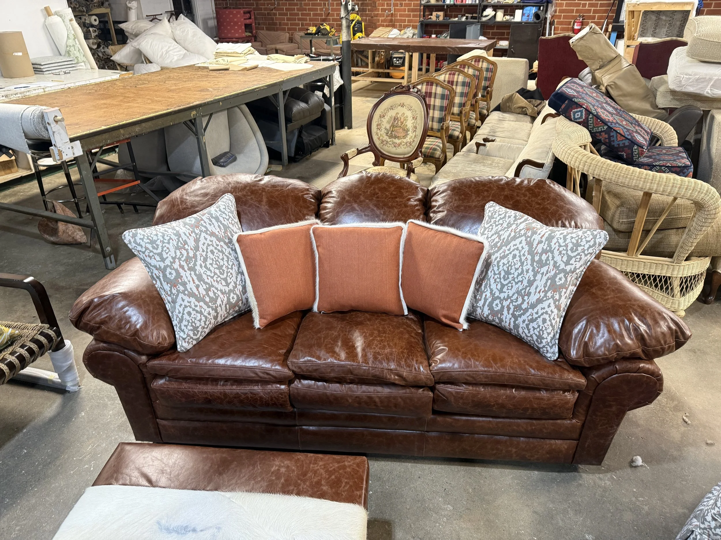 A variety of furniture including a large brown leather couch with decorative pillows, a vintage floral armchair, several plaid and neutral-colored chairs, and a small upholstered ottoman in what appears to be a warehouse or thrift store setting.