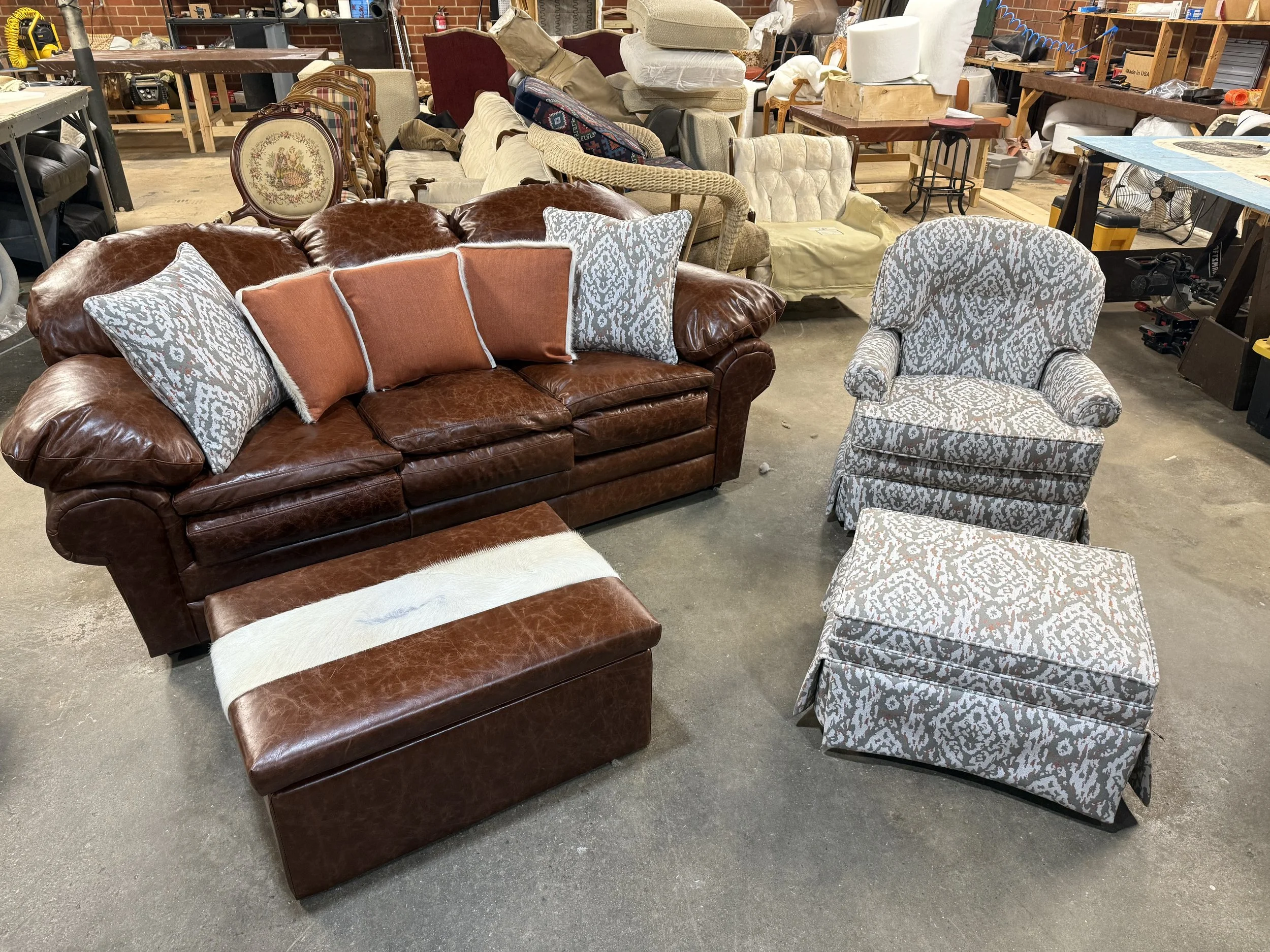 A collection of various used furniture items including a brown leather sofa with pillows, a patterned armchair, a cream vintage sofa, and other chairs, in a warehouse-like setting with a concrete floor and brick walls.