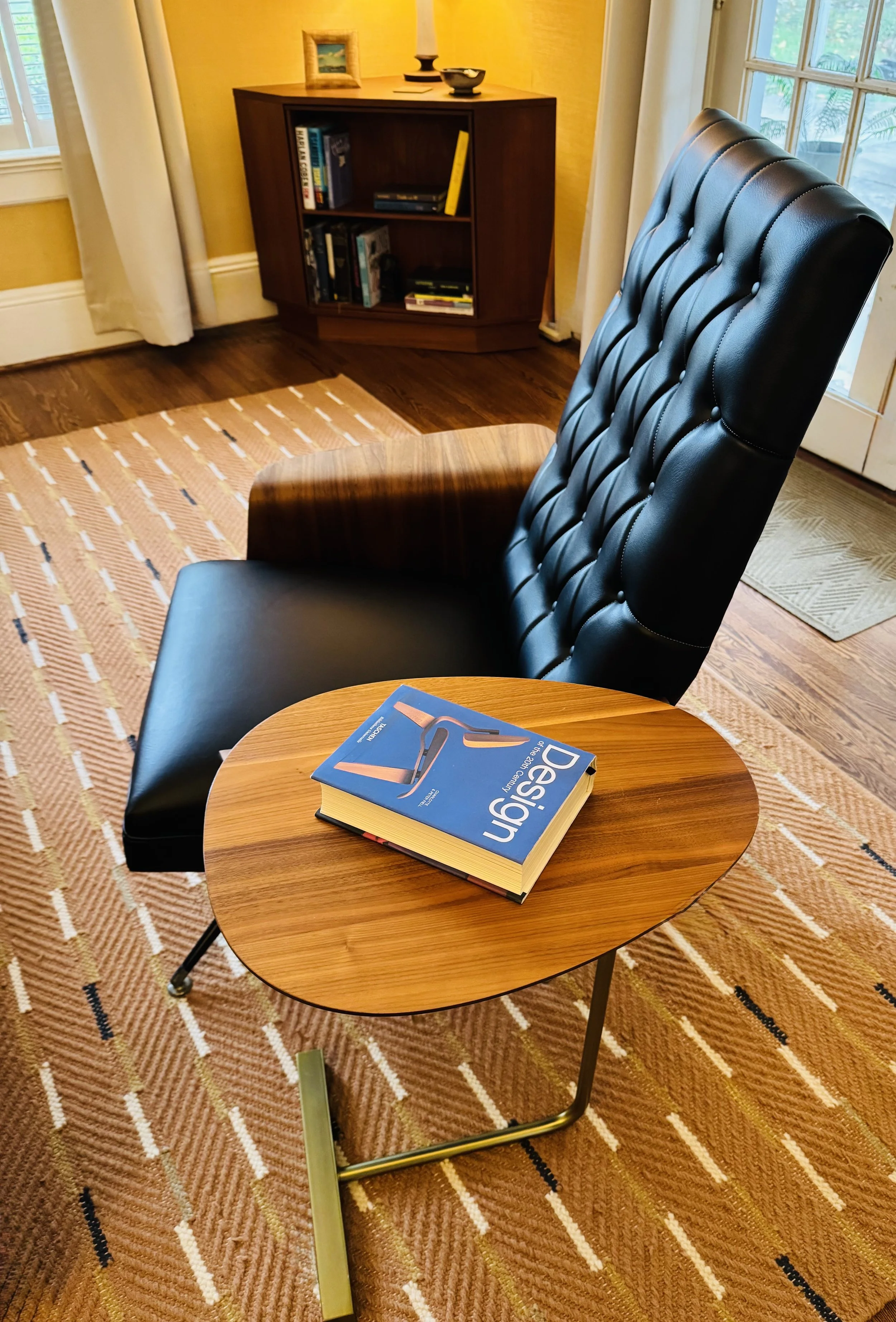 Close-up of a wooden round table with a book on design, a black leather padded chair, a patterned carpet, a bookshelf with books, a window with curtains, and a yellow wall.
