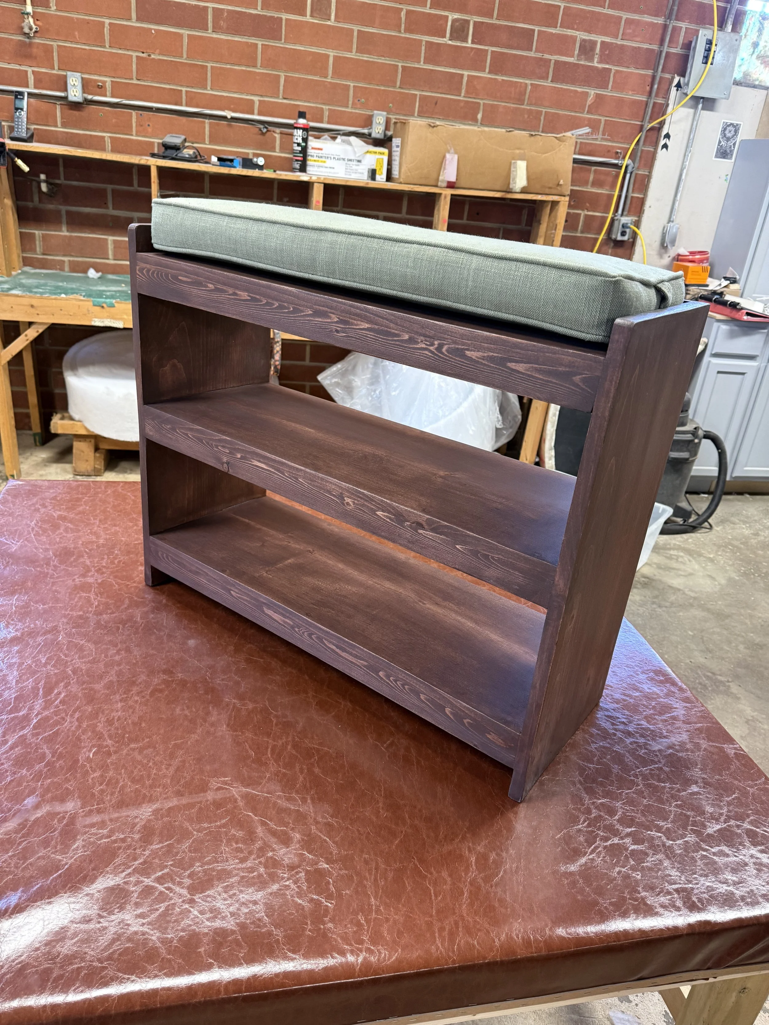 A wooden bookshelf with a green cushioned top in a workshop setting, with a brick wall and various tools and boxes in the background.