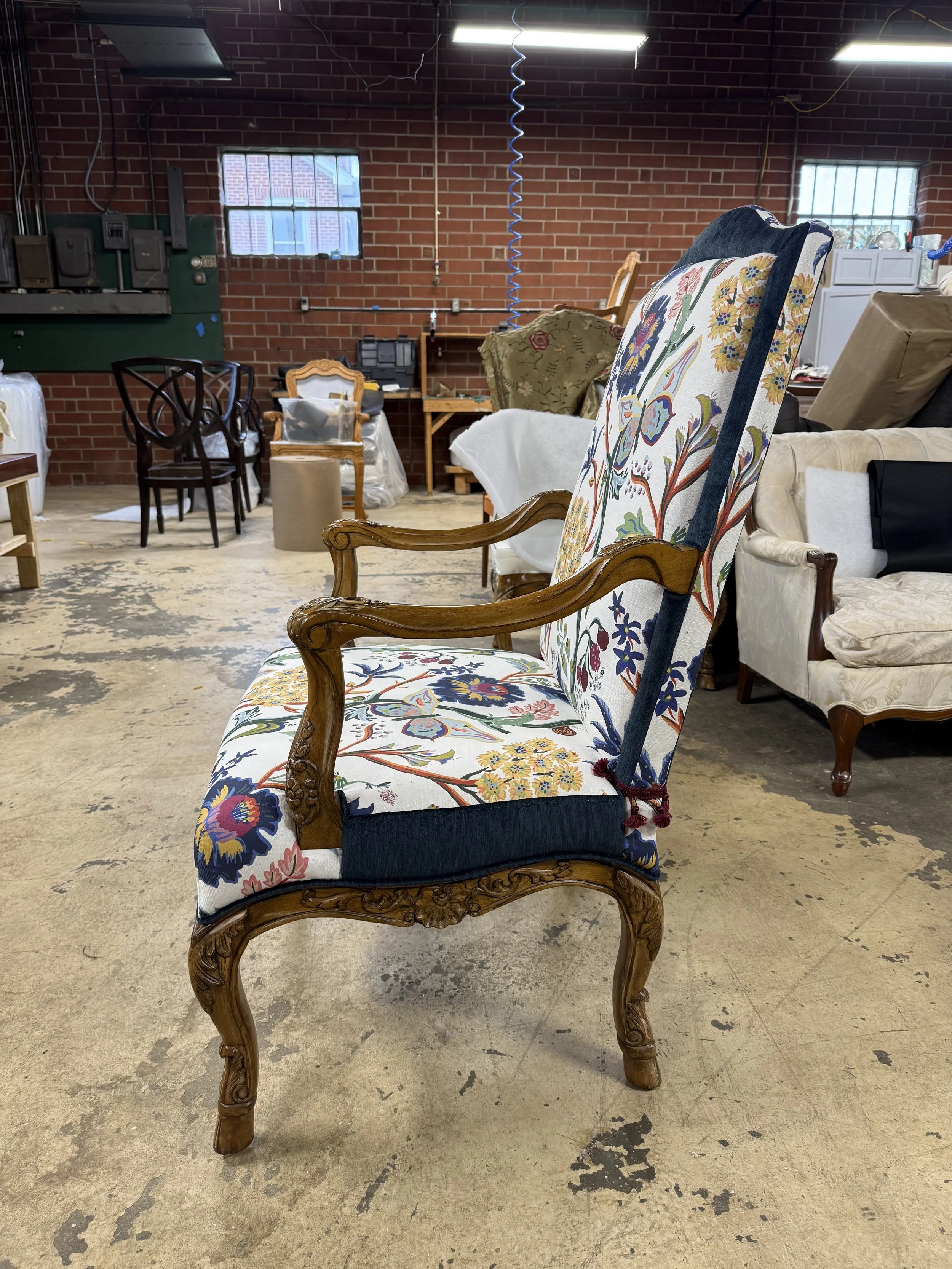 A vintage wooden armchair with floral upholstery in a warehouse or furniture store. Surrounding the chair are other assorted chairs, furniture, and boxes, with exposed brick walls and fluorescent lighting overhead.