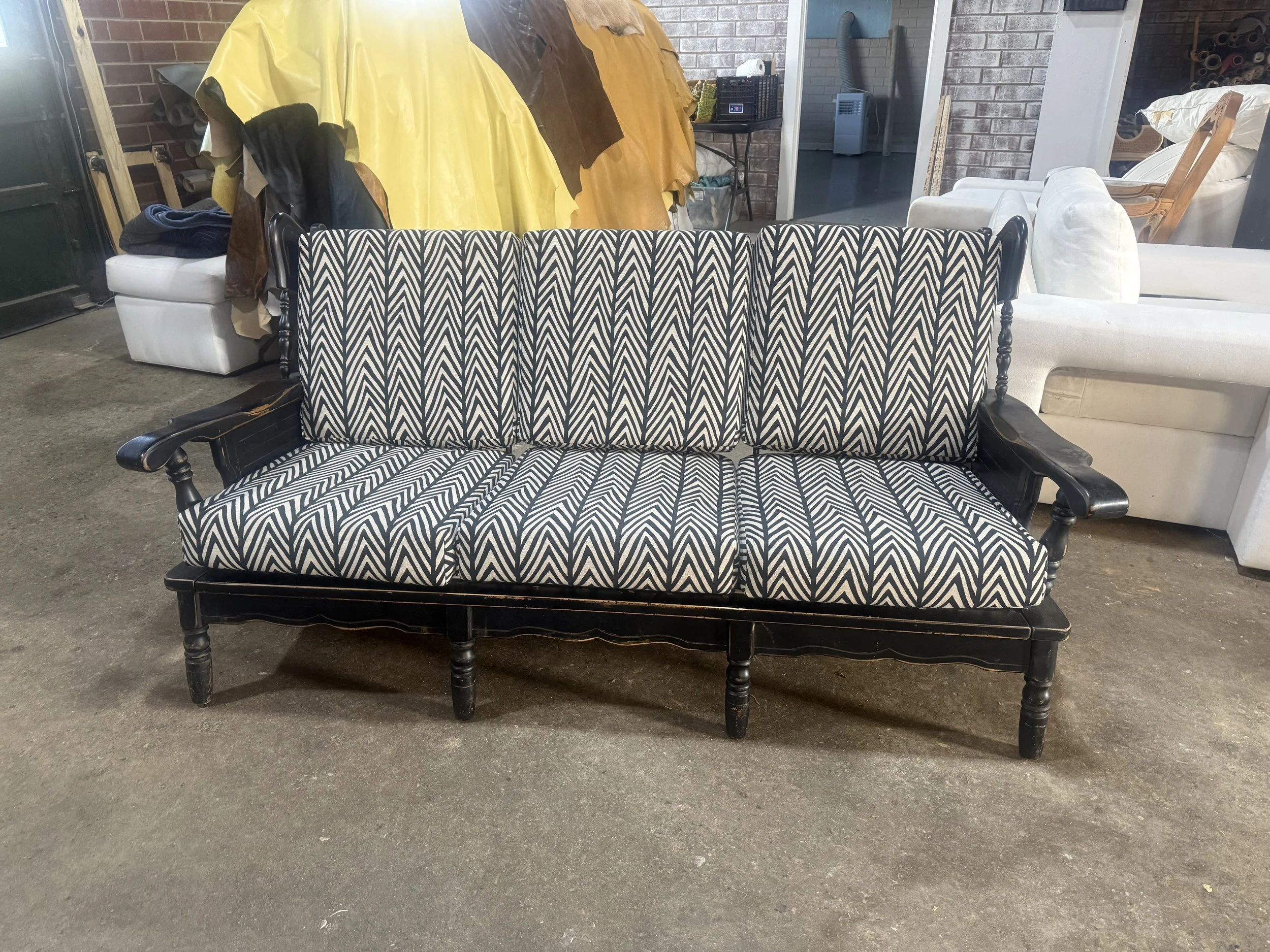 A furniture store with a black and white chevron patterned sofa in the foreground, a white armchair to the right, and a yellow and brown fabric hanging in the background. The store has a concrete floor, exposed brick walls, and various furniture item