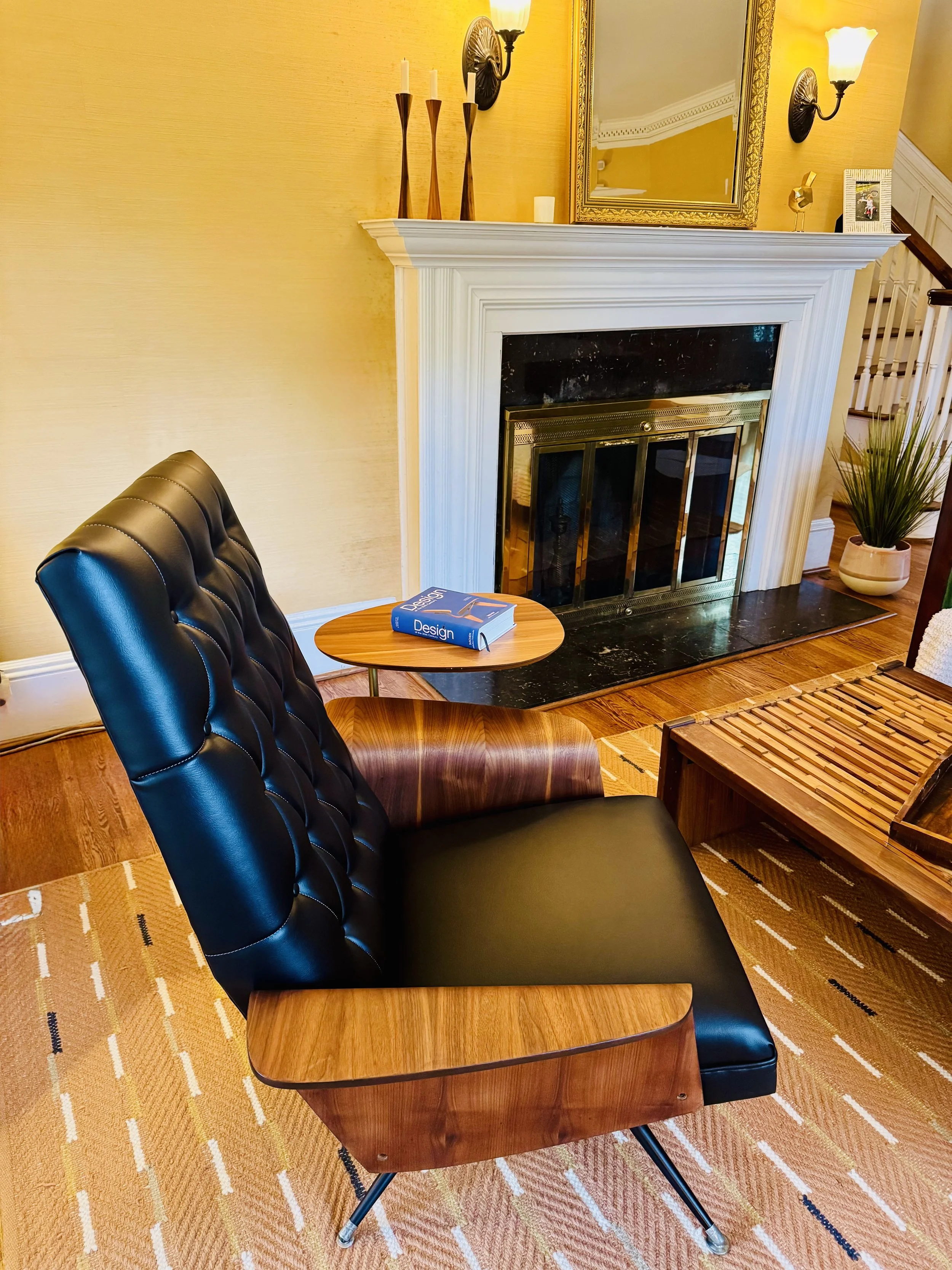 A cozy living room area with a black leather and wood armchair, a small wooden side table with a book titled 'Design,' a fireplace with a black and gold finish, a large mirror above the fireplace, wall sconces with warm lighting, a potted plant in a 