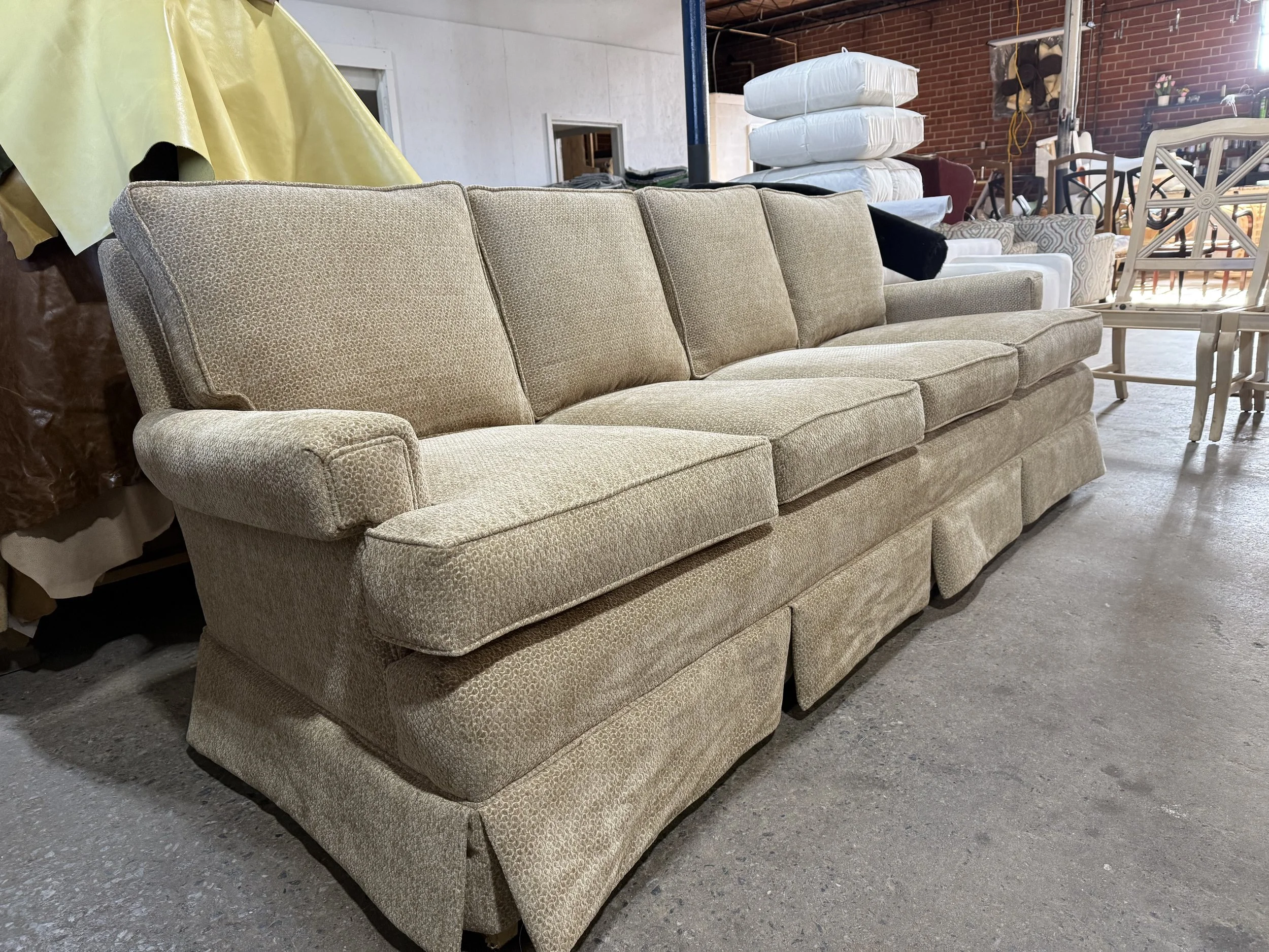 A beige upholstered sofa with a textured fabric sits in a cluttered basement or storage area with various furniture and items stacked behind and beside it.