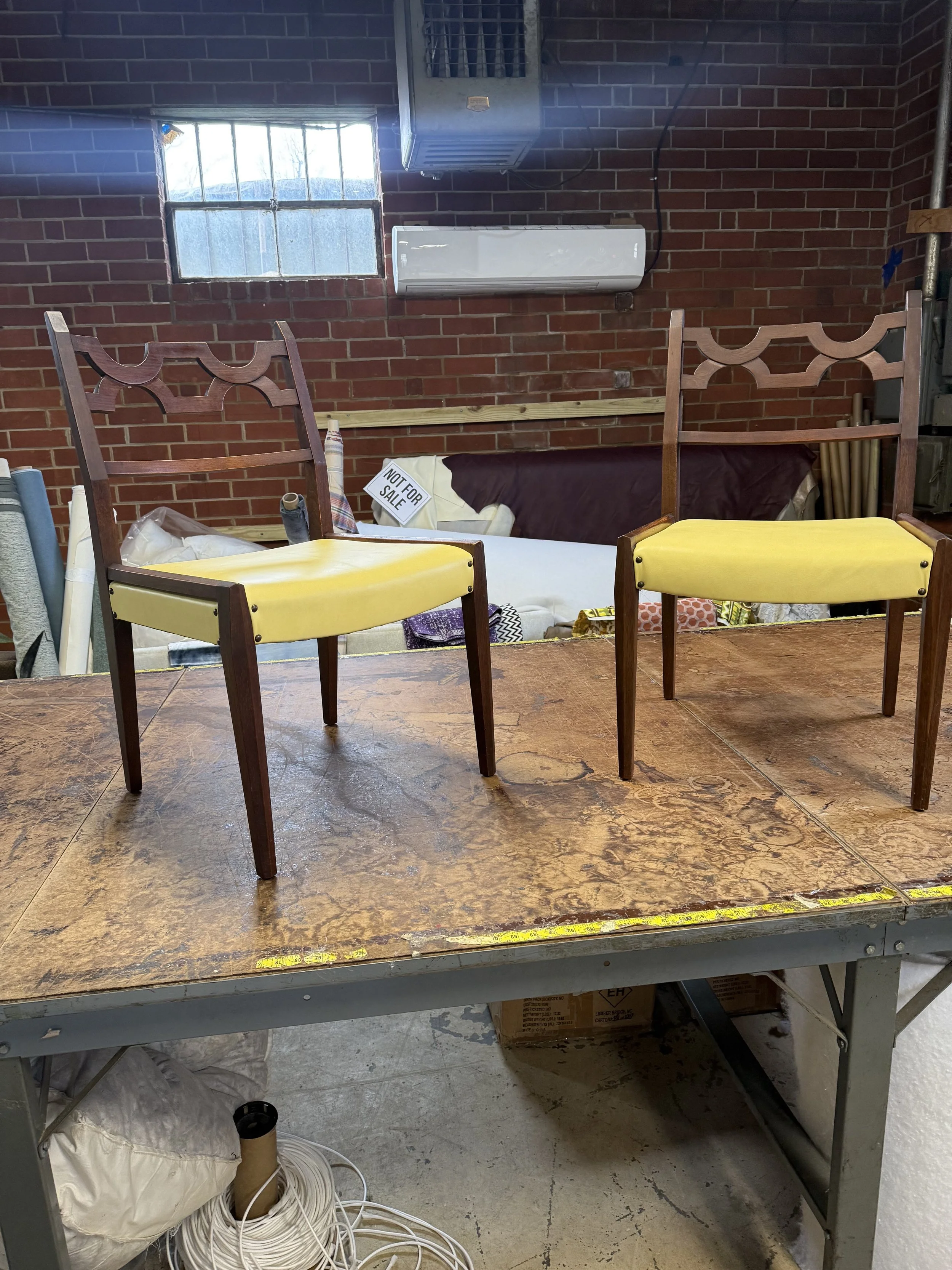 The image shows a workbench with two chairs with yellow seats in front. Behind the workbench, there's a red brick wall with a small window. Various items like fabric rolls, a sign reading 'NOT FOR SALE,' and coiled white extension cords are on or bel