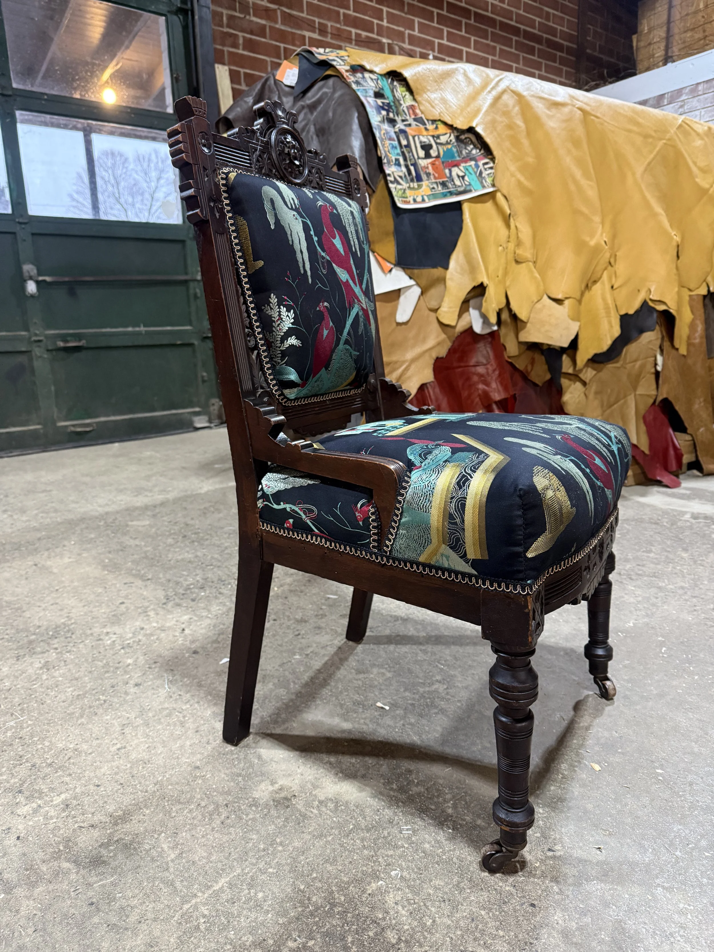 An antique wooden chair with embroidered fabric on the seat and backrest, placed in an industrial-style setting with a concrete floor, brick wall, and various fabrics and leather stored on a table in the background.