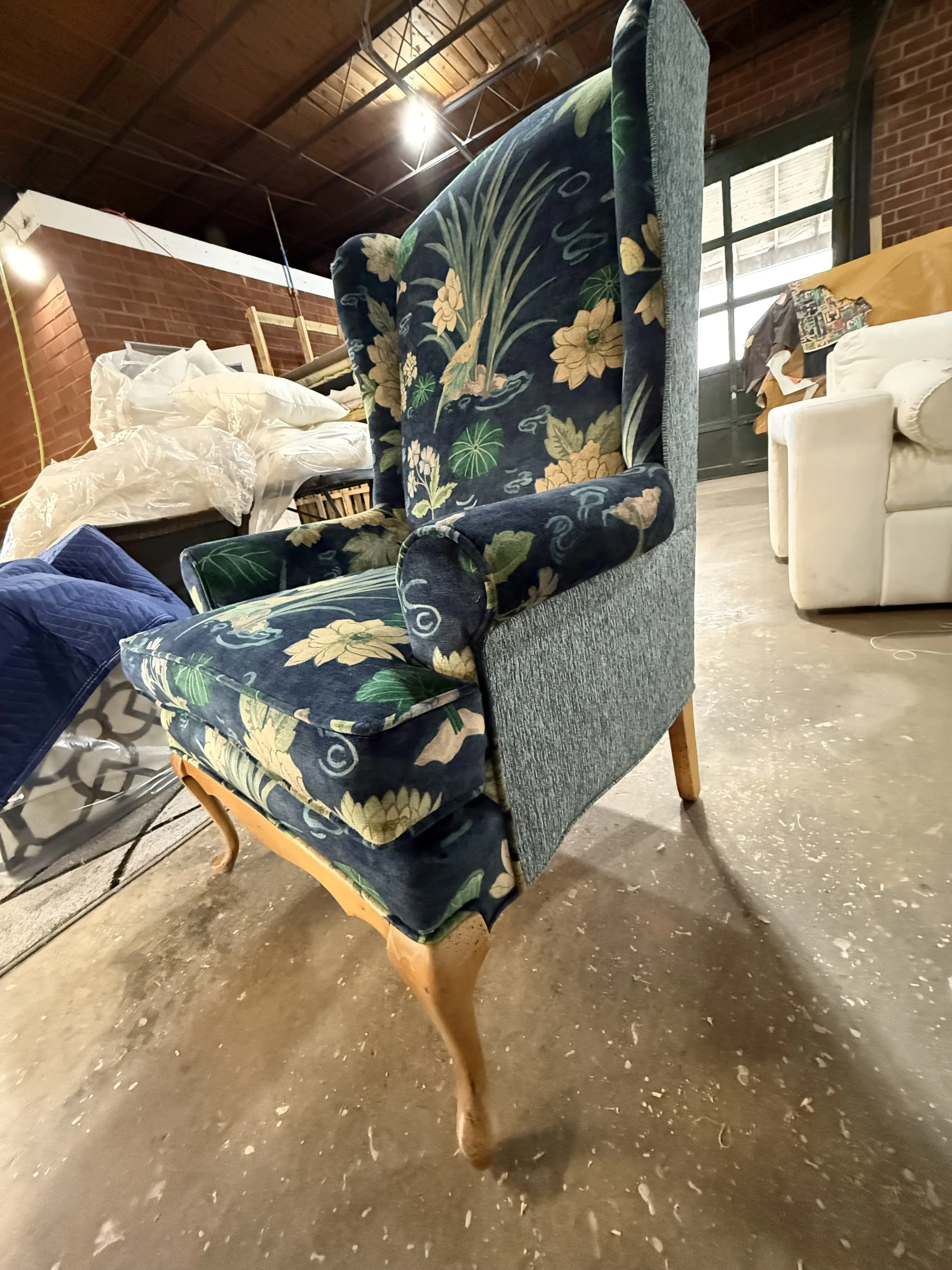 A vintage armchair with a floral pattern on its upholstery, standing on a concrete floor in an industrial-style room with brick walls and large windows.