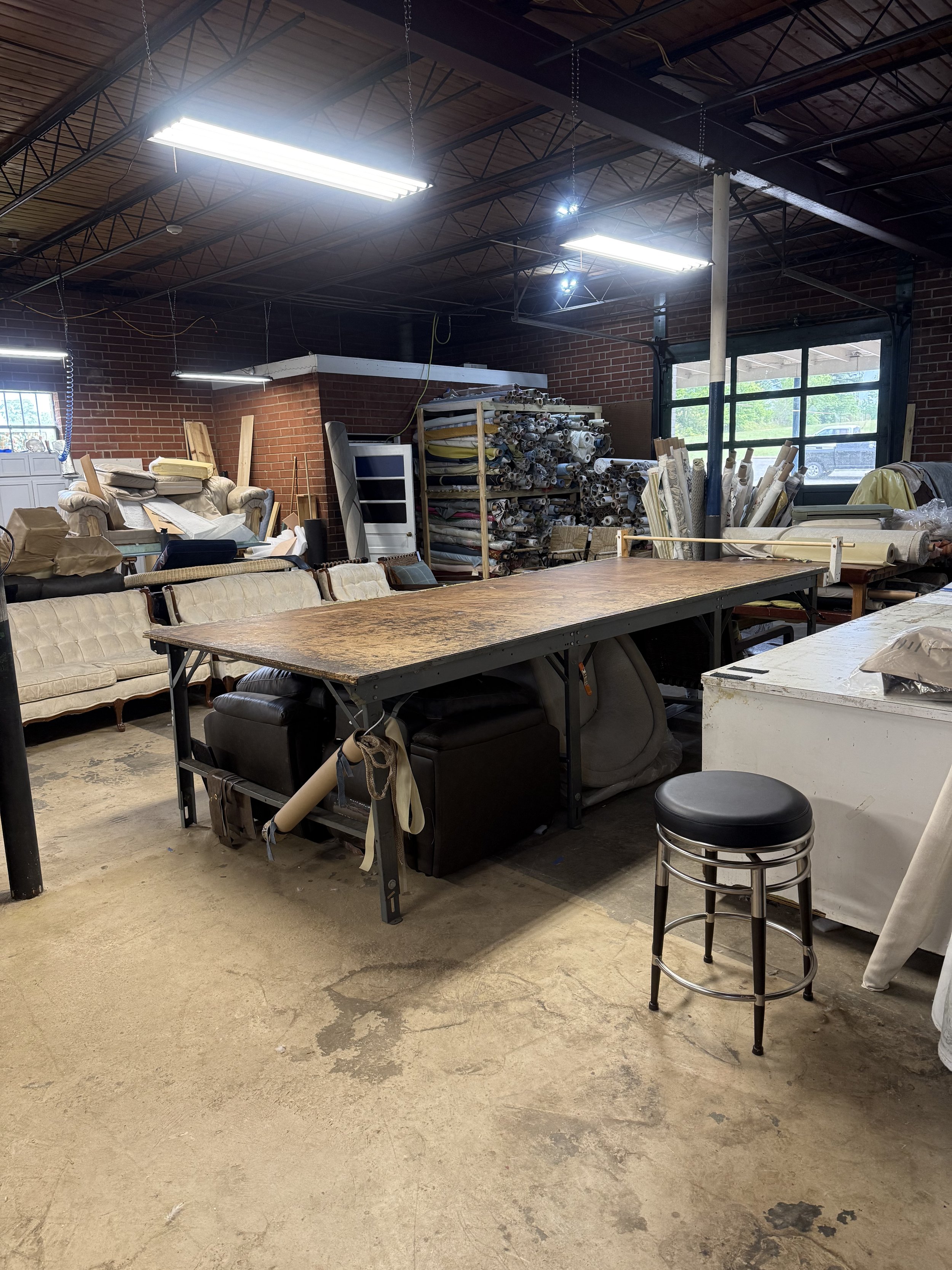 Interior of a workshop with a long wooden table, a vintage couch, and shelves filled with rolled materials. Bright fluorescent lights on a high ceiling with brick walls and a large window showing a parking lot outside.