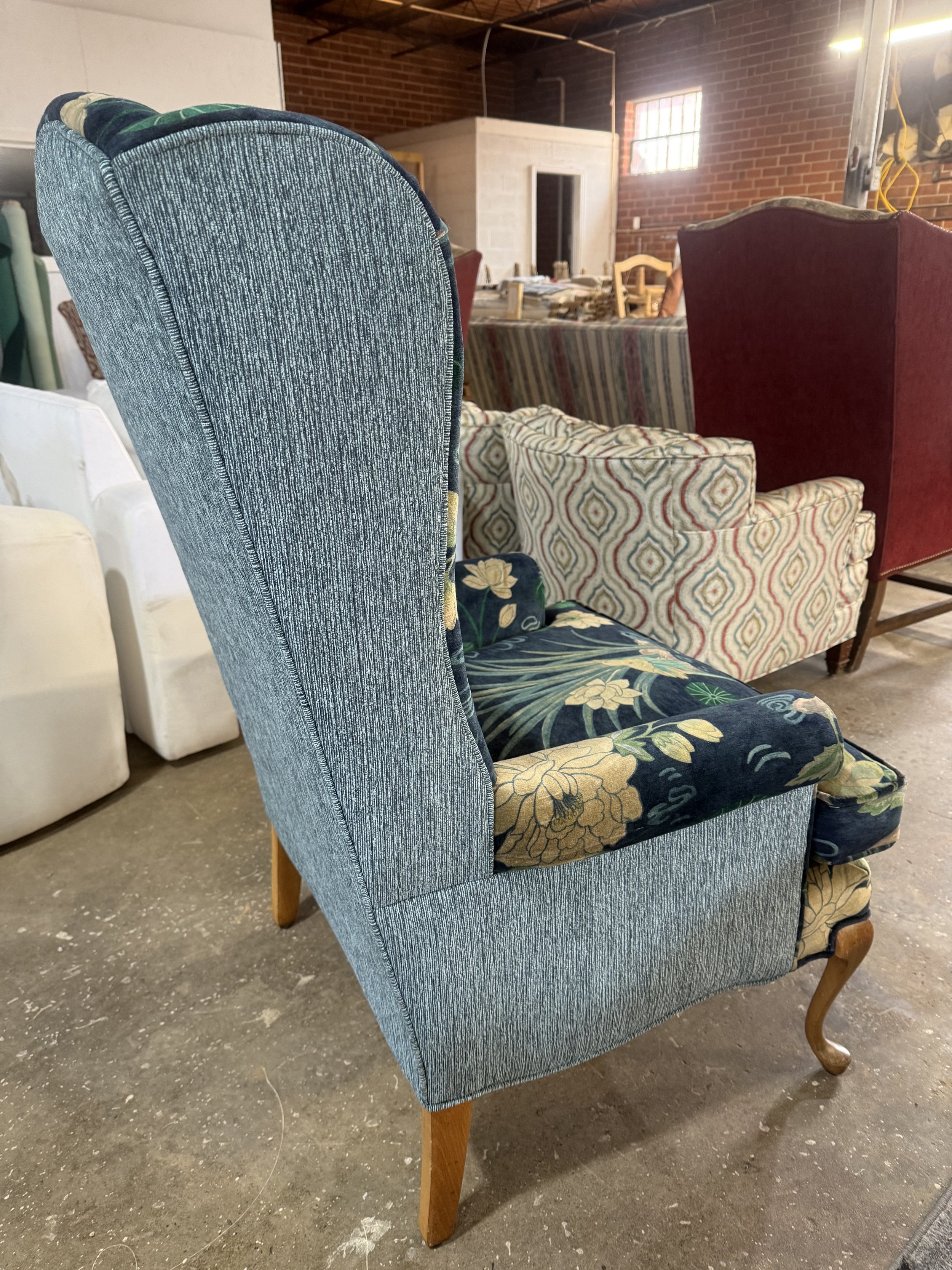 A vintage high-back upholstered armchair with floral fabric on the seat and a light blue fabric on the backrest, wooden legs, in an industrial-style room with brick walls and tables and chairs in the background.