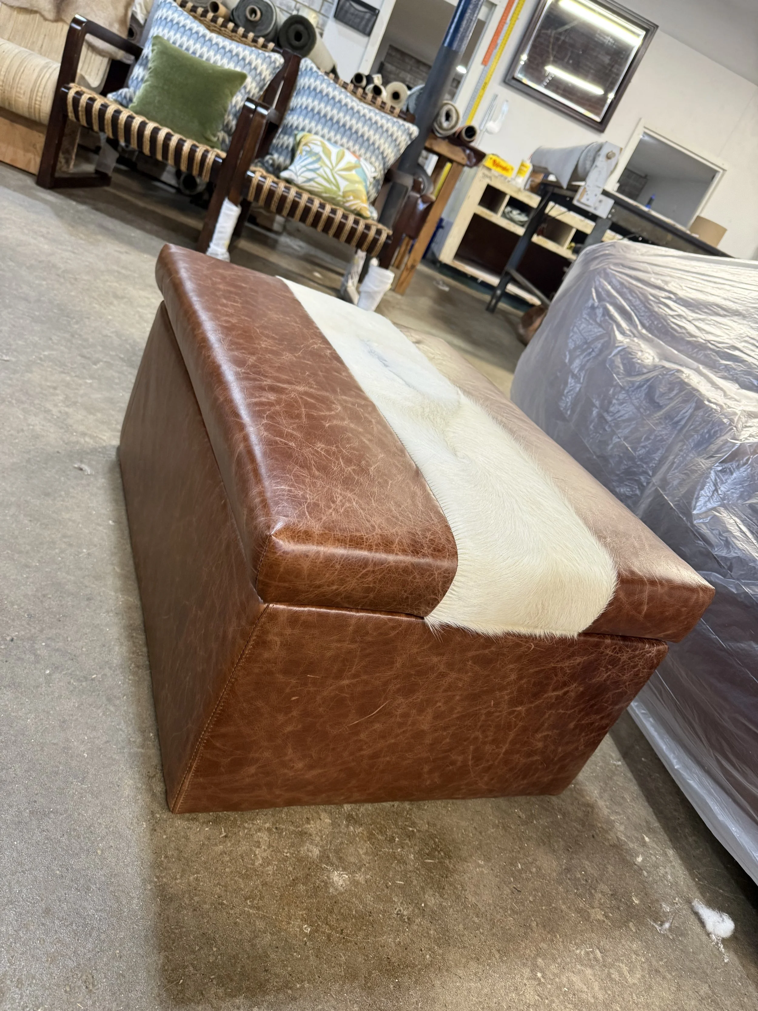 A brown leather ottoman positioned on a concrete floor in front of a sofa, with a white and beige fur throw draped over part of the ottoman. In the background, there are patterned armchairs, shelves, and a workbench with various items.