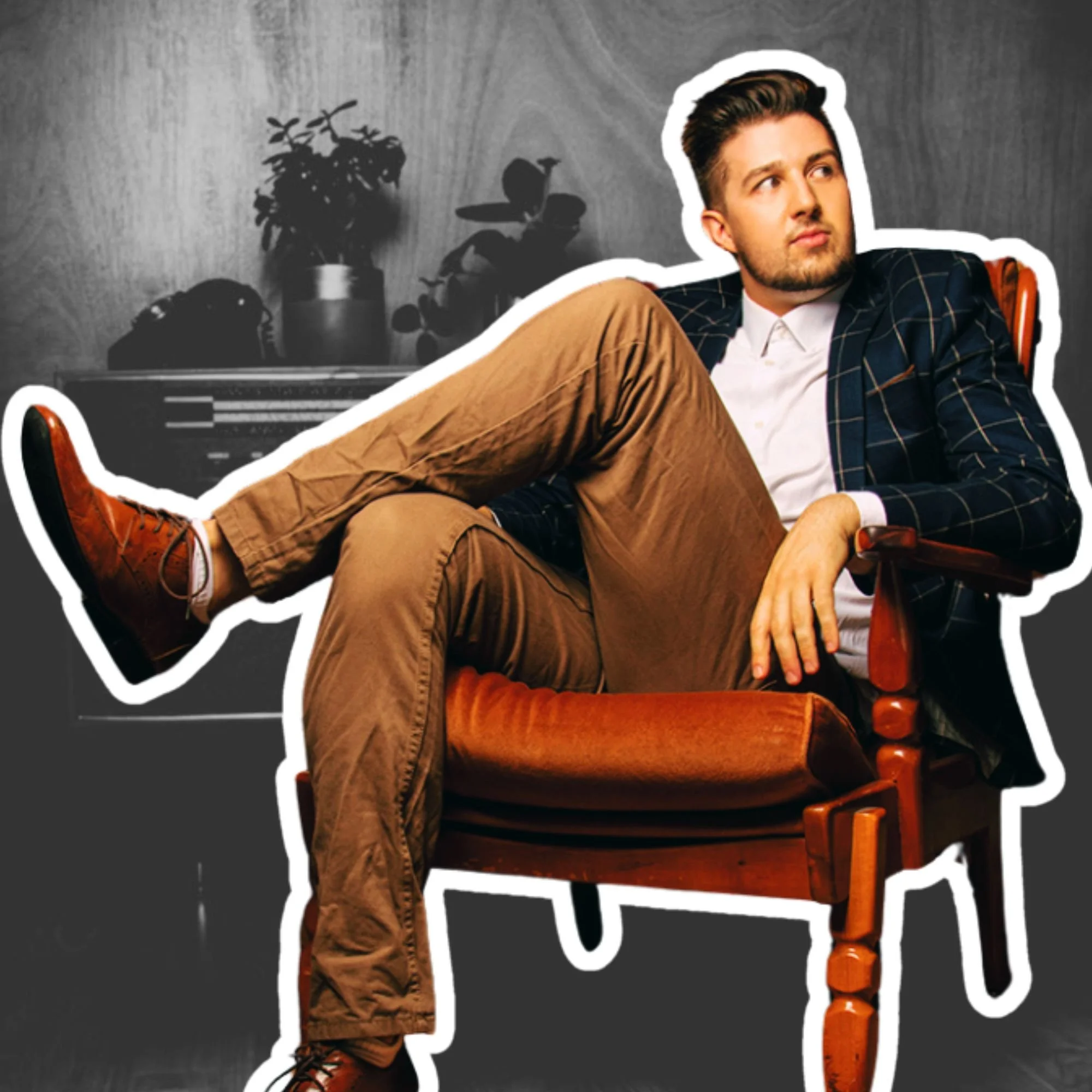 A young man with dark hair and a beard, wearing a white shirt, dark checkered blazer, and brown pants, sitting casually in a wooden armchair with one leg resting on the armrest, looking to the side. Kai Mann-Robertson