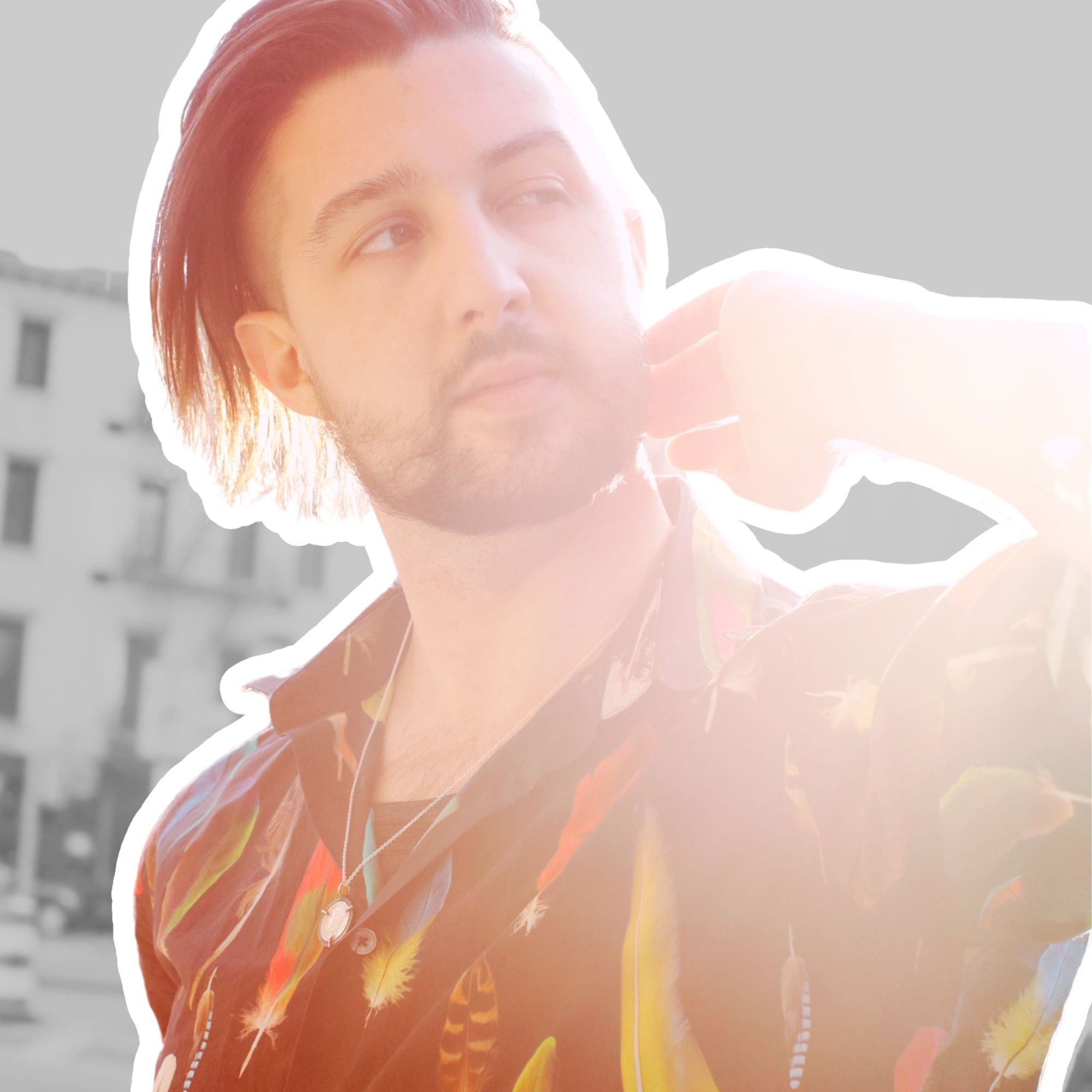 Kai Mann-Robertson Young man with medium-length hair, facial hair, and wearing a colorful shirt, looking to the side with sunlight shining on him, standing outdoors in front of a building.