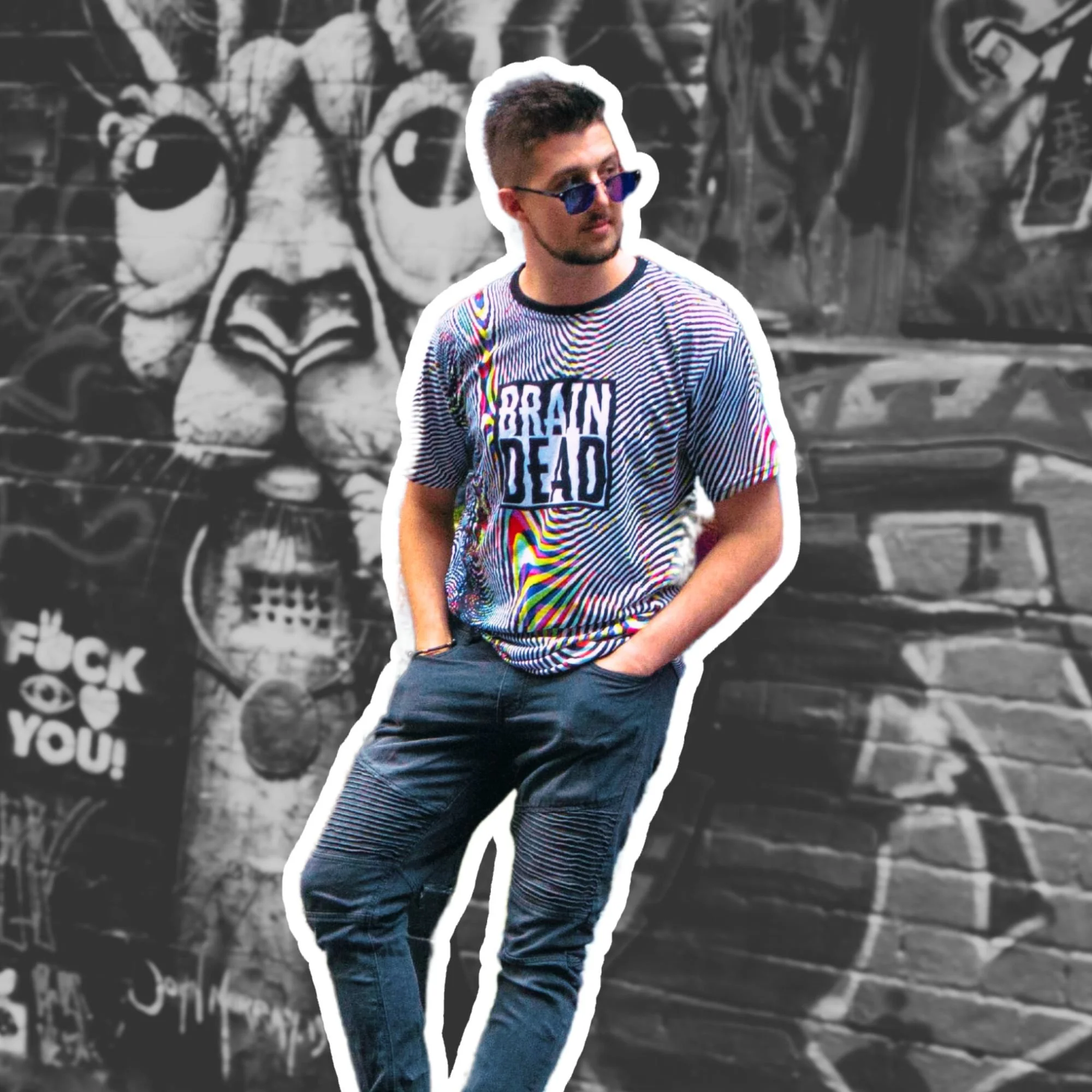 A young man Kai Mann-Robertson with sunglasses and a graphic t-shirt that says 'BRAIN DEAD' standing against a graffiti-covered brick wall.