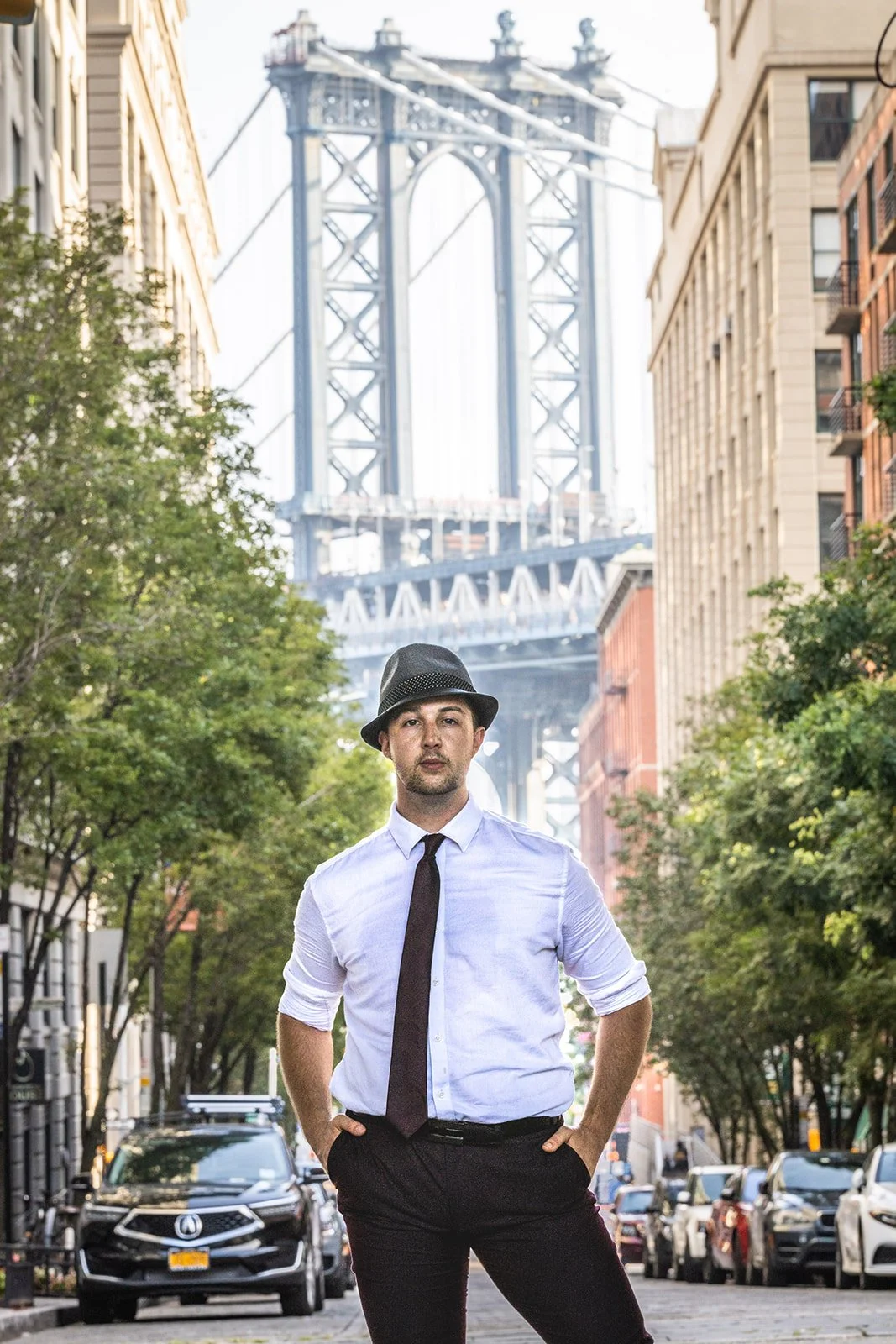 Kai Mann-Robertson wearing a white shirt, black tie, and gray fedora hat standing on a city street, with the Brooklyn Bridge in the background.