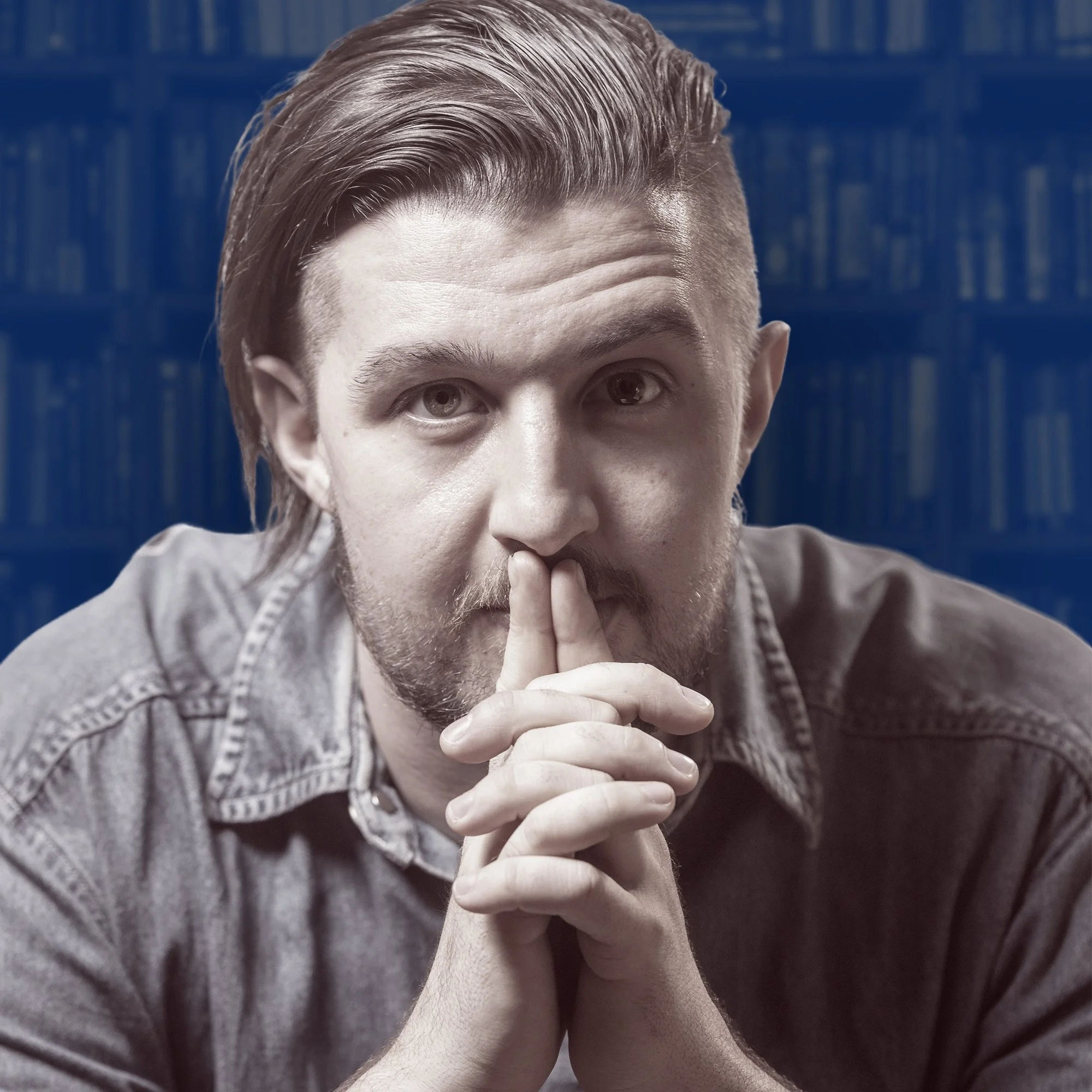 Kai Mann-Robertson with styled hair and a beard holding his hands together near his face, with a thumb and index finger crossed in a contemplative pose, against a background of a bookcase filled with books.