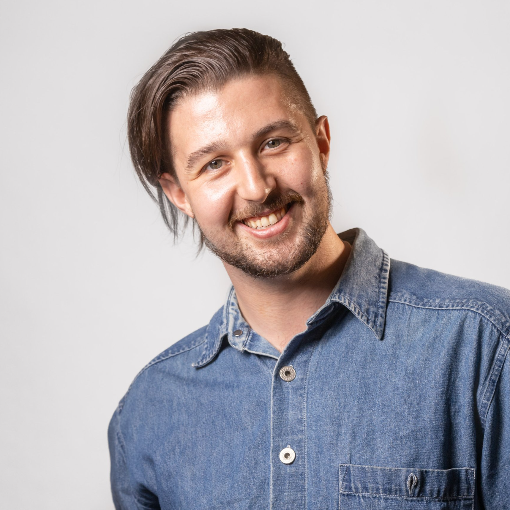 Smiling man with medium-length hair and beard wearing a denim shirt against a light background. Kai Mann-Robertson