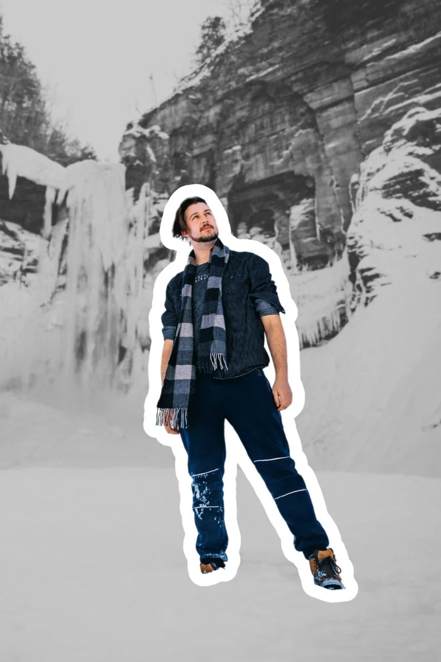 A man standing outdoors in a snowy landscape with icy waterfalls and rocks in the background, with a black and gray scarf, dark jacket, and snow boots.