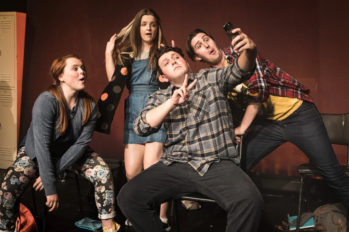 Five young people taking a selfie together on a stage in a school setting.