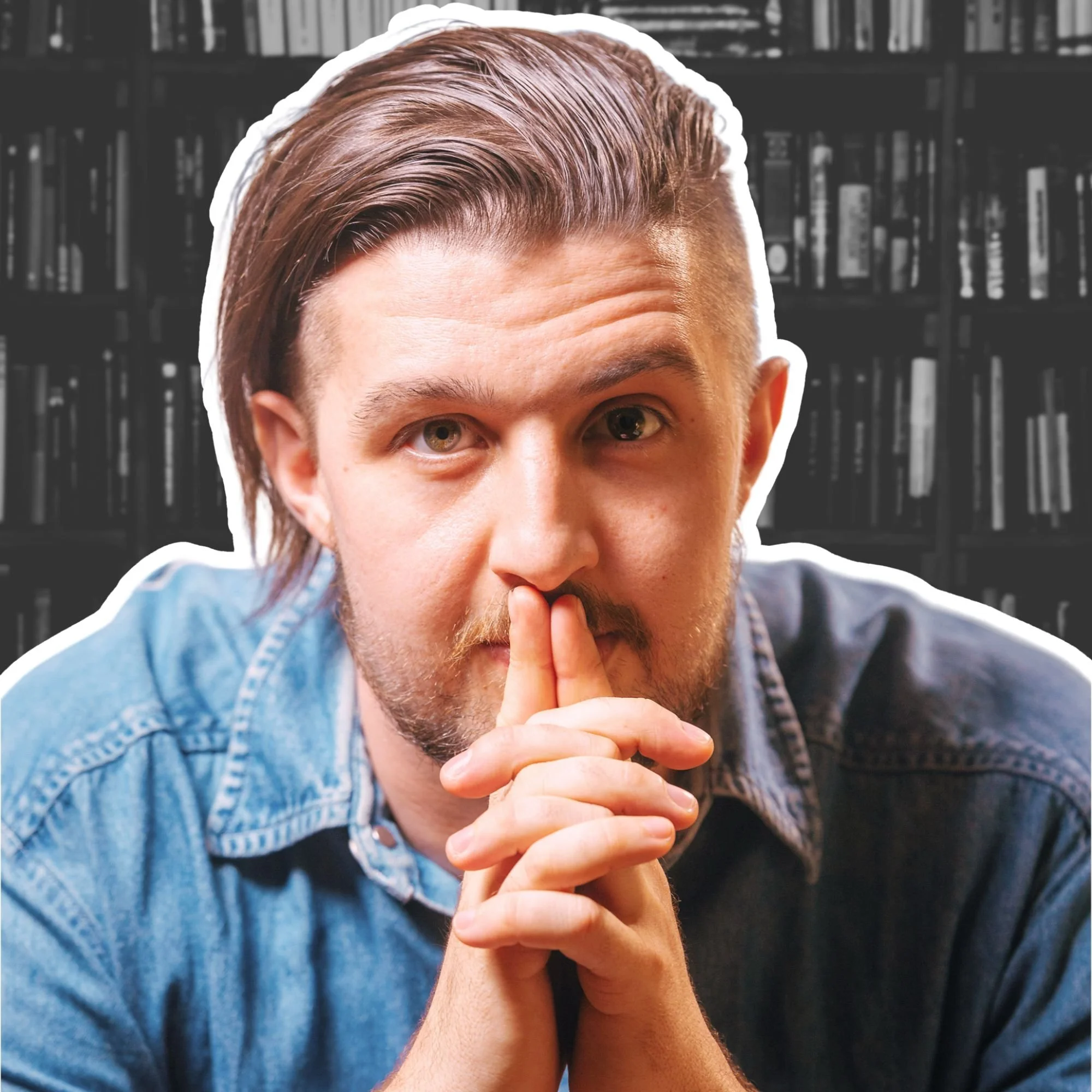 Kai Mann-Robertson A man with medium-length brown hair and a beard, wearing a denim shirt, sitting in front of a bookshelf, holding his hands in front of his face with his index fingers touching his lips, looking directly at the camera.