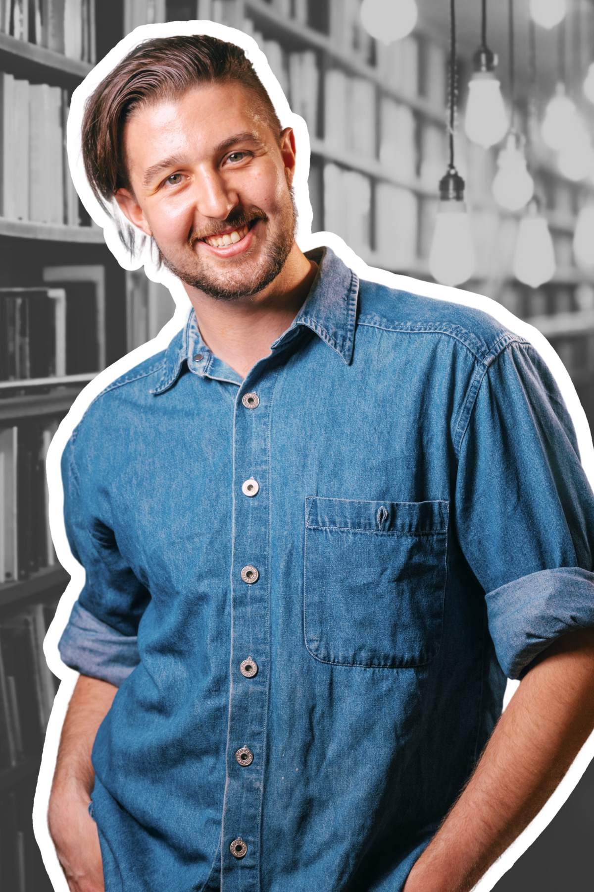 Kai Mann-Robertson Happy young man with a beard and mustache, wearing a denim shirt, smiling with hands in pockets, standing in a library with shelves and hanging lights in the background.