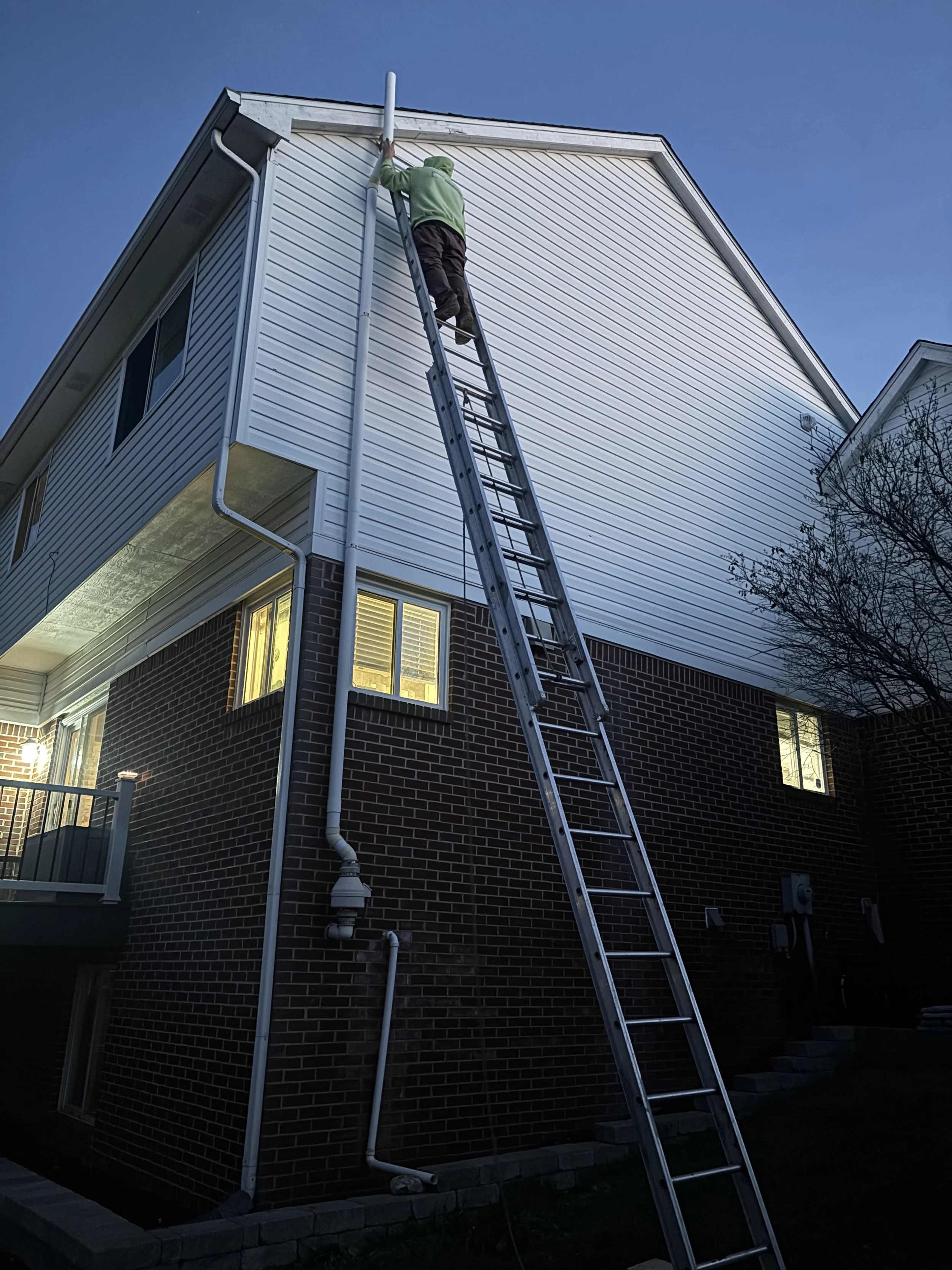 A person in a green hoodie and black pants standing on a tall extension ladder, working on the upper exterior of a house with white siding, during dusk or evening.