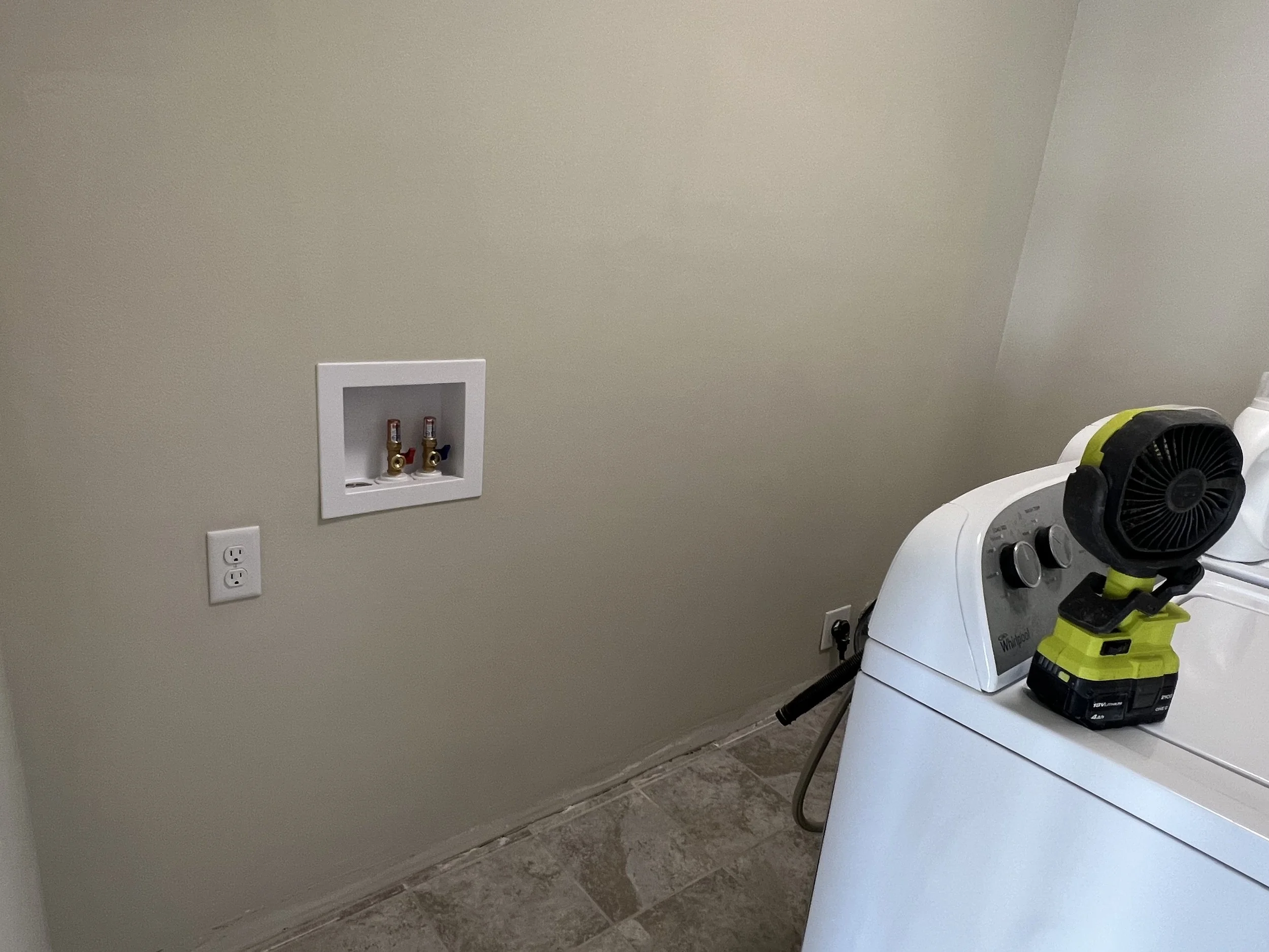 A laundry room with a washing machine, a cordless drill, and a portable fan on top of the washing machine. There is an electrical outlet and a boxed set of water supply valves on the wall.